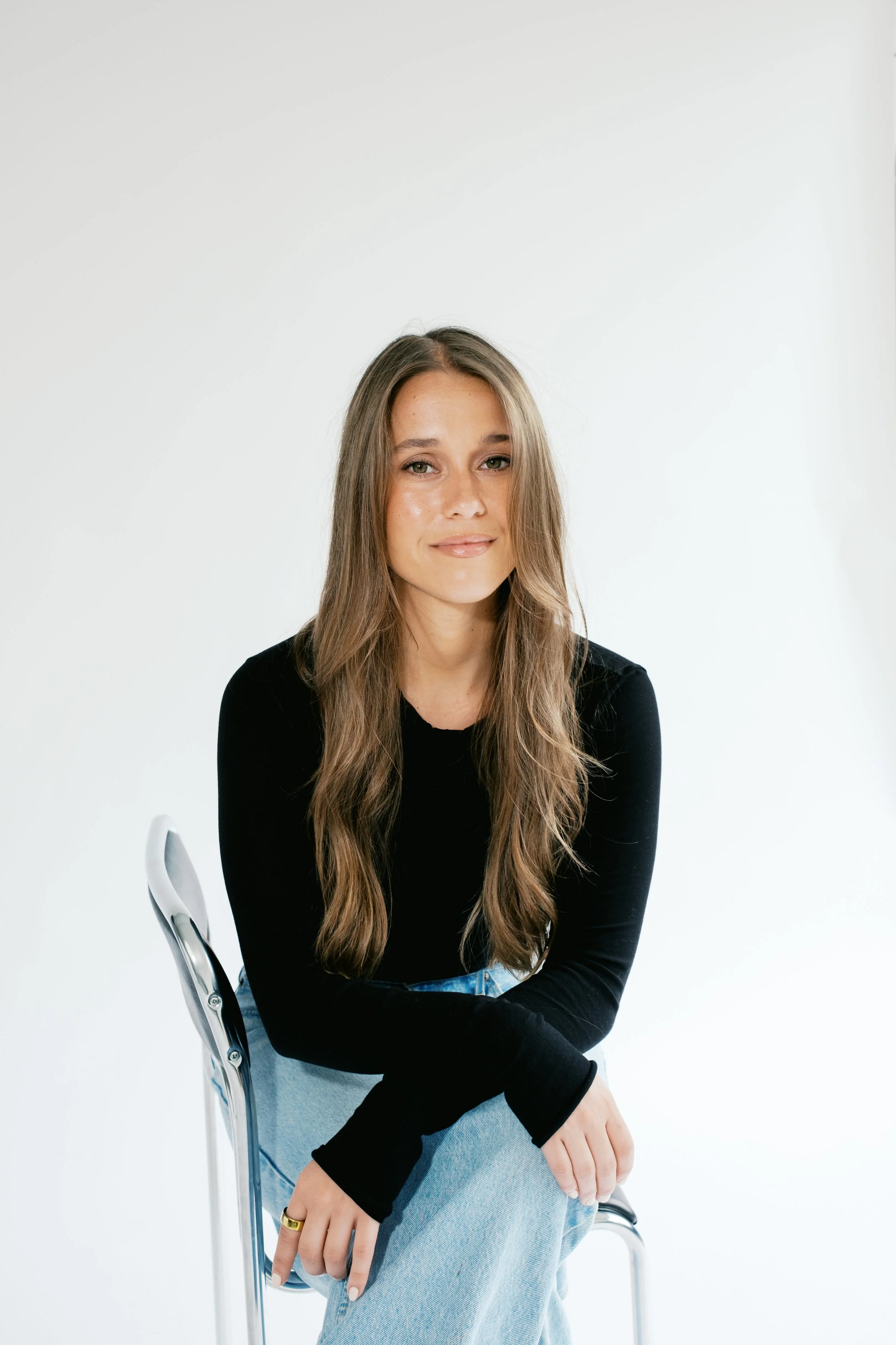 Young woman with long brown hair wearing a black top and light blue jeans, sitting on a silver chair against a plain white background.