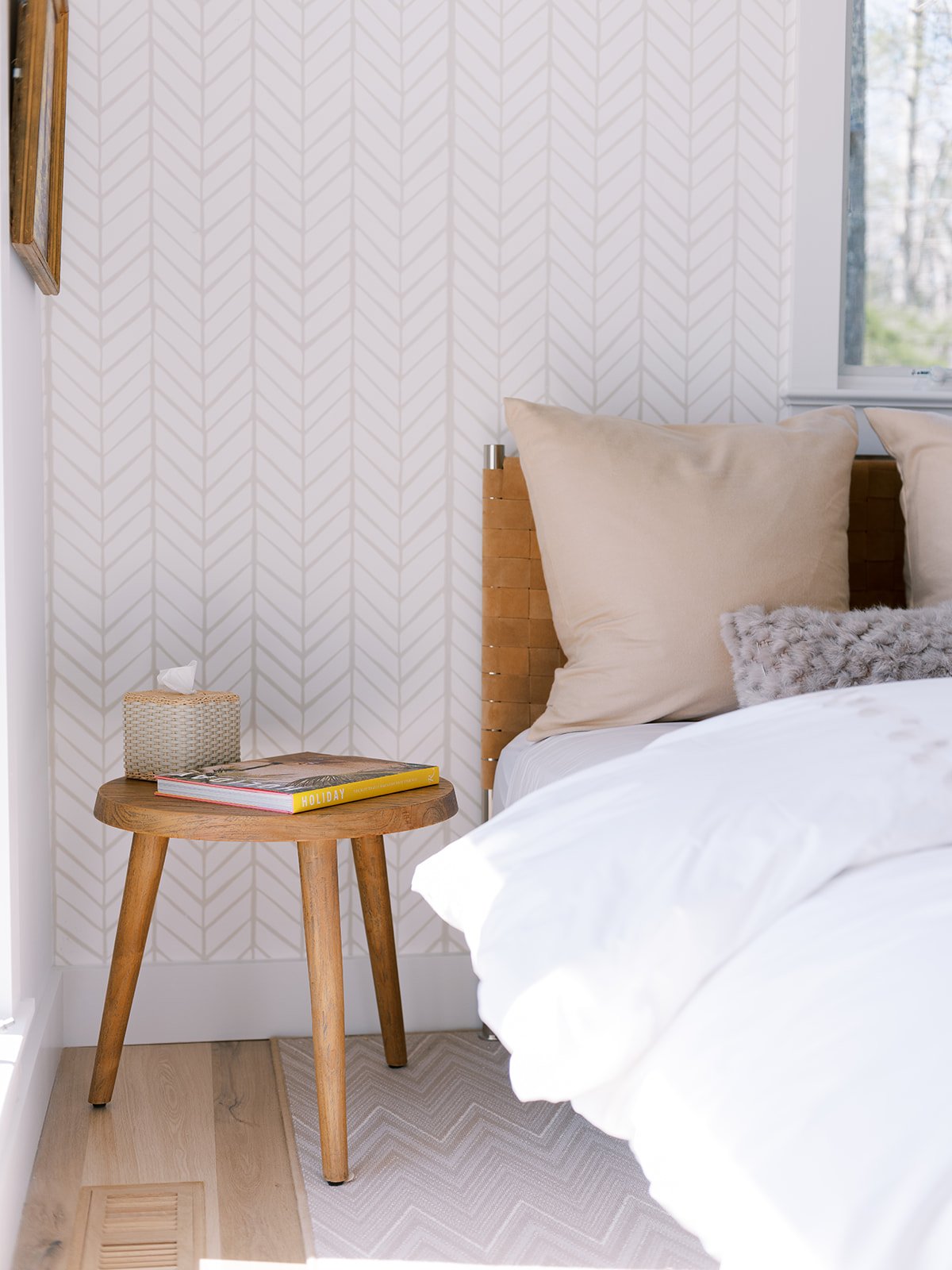 A cozy bedroom corner with a bed that has beige pillows and a textured throw, a small round wooden side table holding a tissue box and a magazine titled 'Holiday,' and a patterned wallpapered wall in the background with a window showing trees outside.