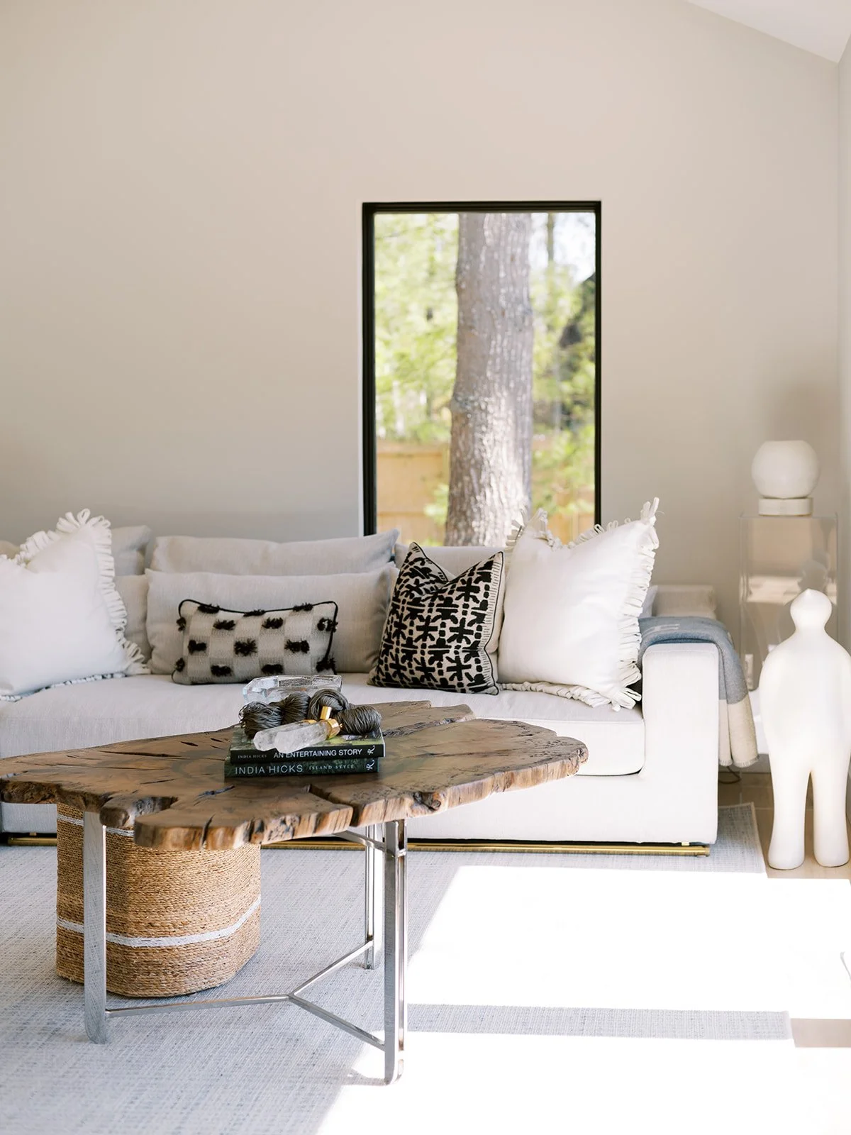 Living room with a white sofa, black and white throw pillows, a rustic wooden coffee table, a large window showing trees outside, and decorative items including a white dog-shaped sculpture and a table lamp.