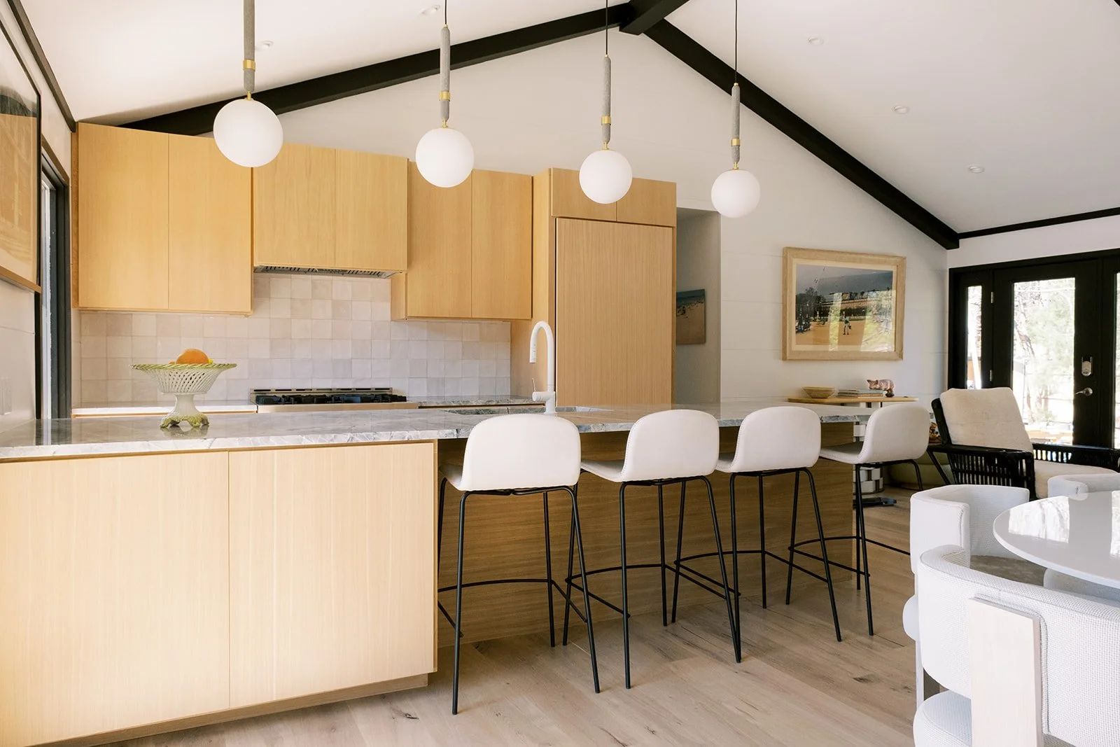 Modern kitchen with light wood cabinets, marble countertops, and five white barstools at the island. Hanging globe lights, framed landscape art, and a seating area with white chairs are visible.