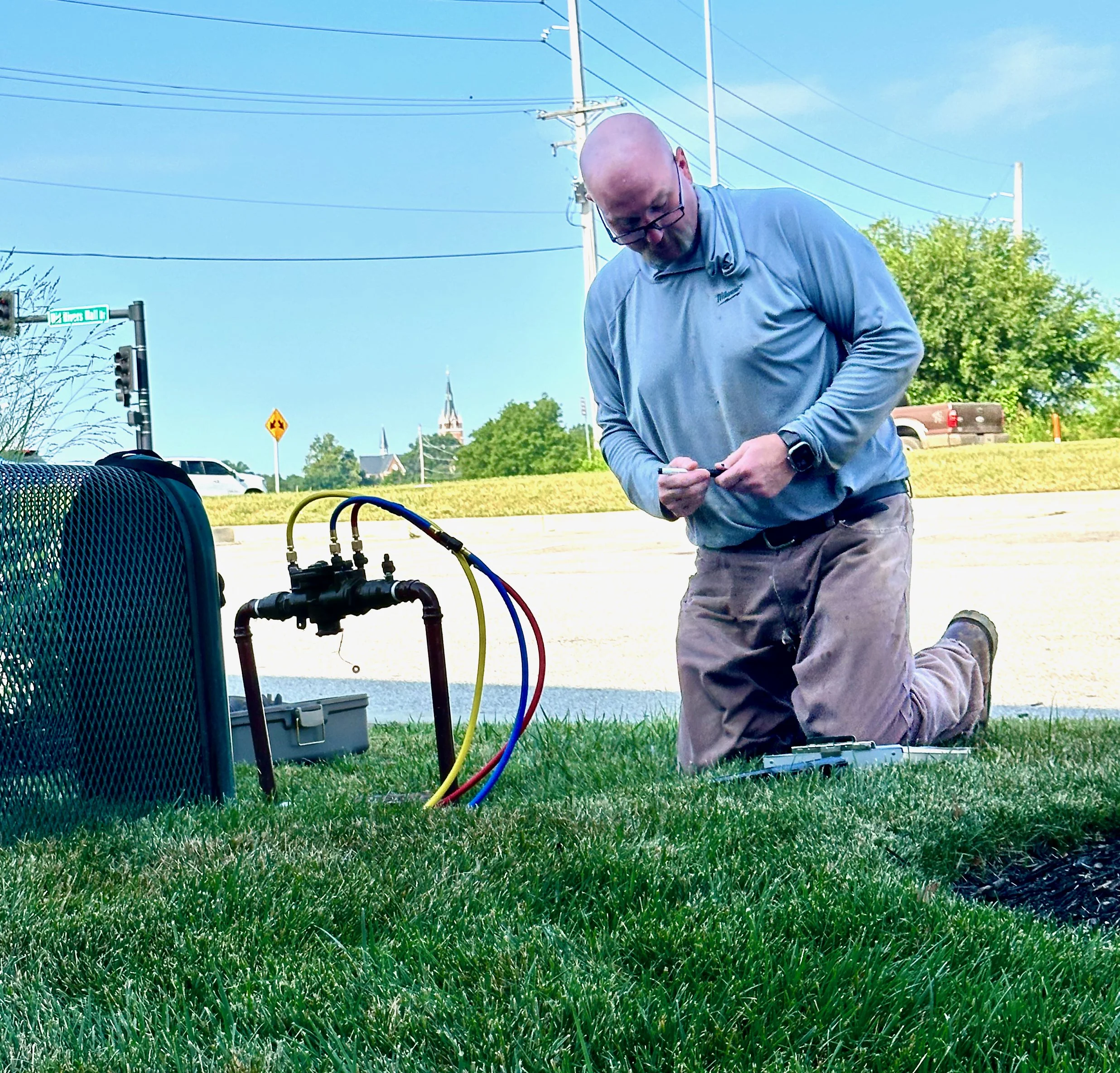 A man kneeling on the grass outdoors, working with electronic equipment connected by colorful cables, with trees, a street sign, cars, and a church steeple in the background.