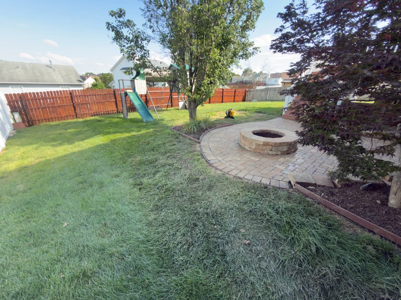 Backyard with green lawn, a wooden fence, a tree, a small slide, a fire pit, and a paver patio.