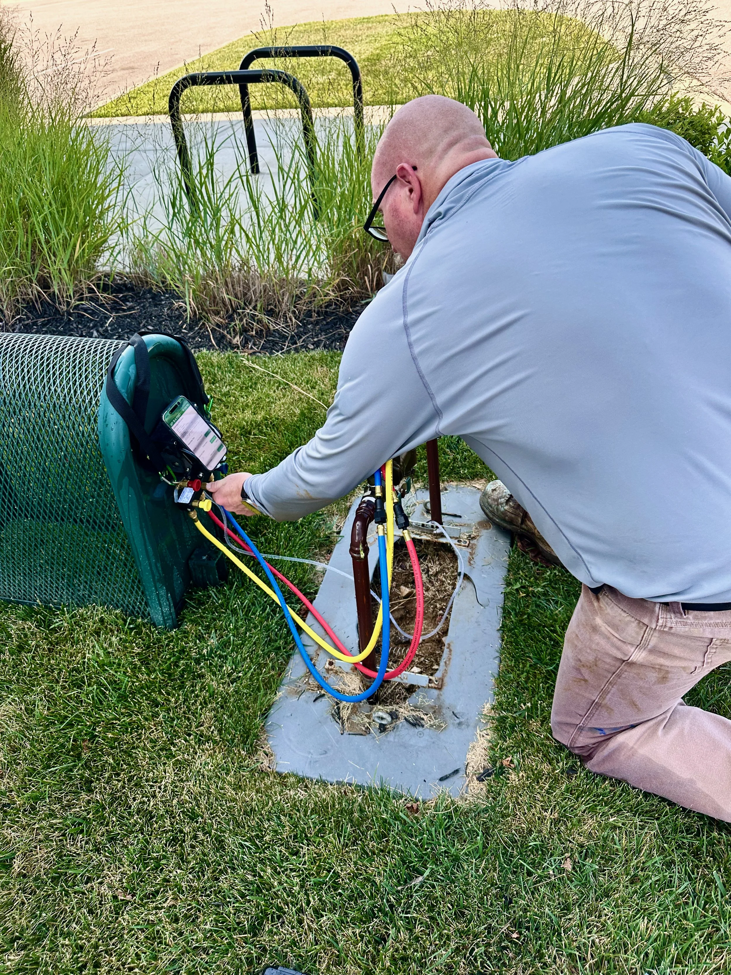 A man kneeling on the grass working with electrical equipment near an outdoor utility box. The box has wires connected to it, and the man appears to be doing maintenance or repair work. There is a green bag and a smartphone nearby, and a black railing is in the background.