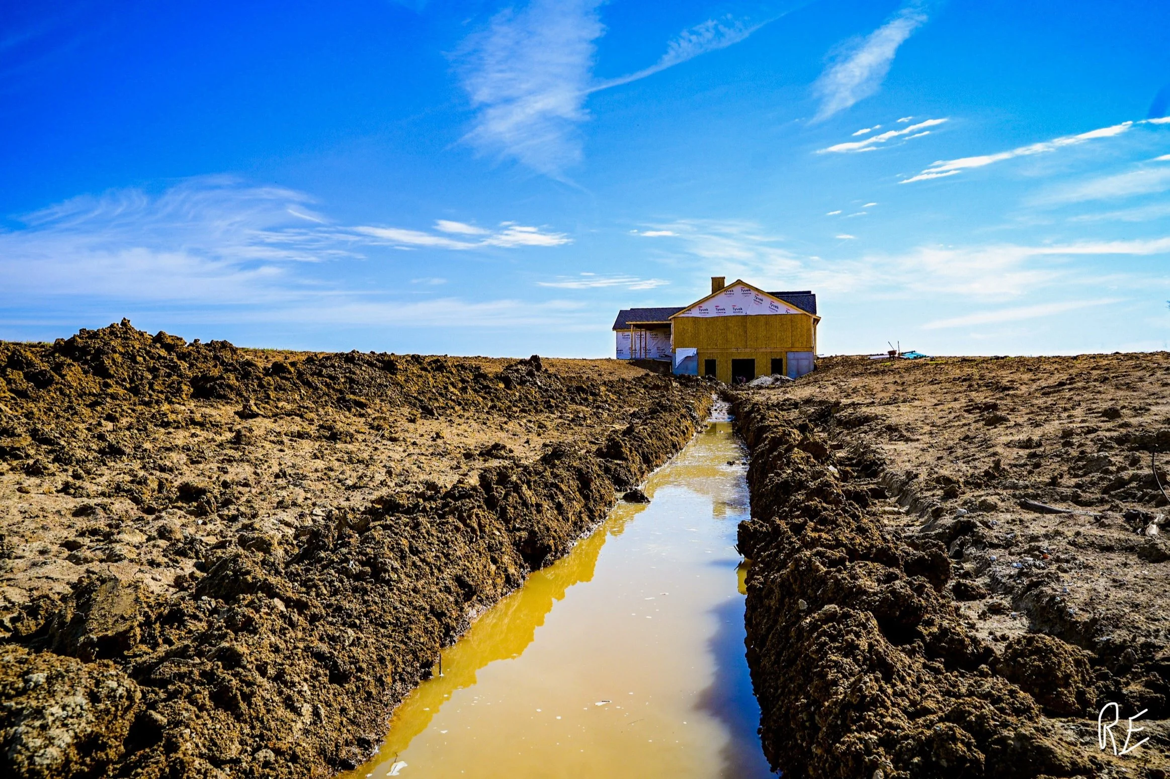 A house under construction on a dirt lot, with a narrow trench filled with water leading towards it, under a partly cloudy blue sky.