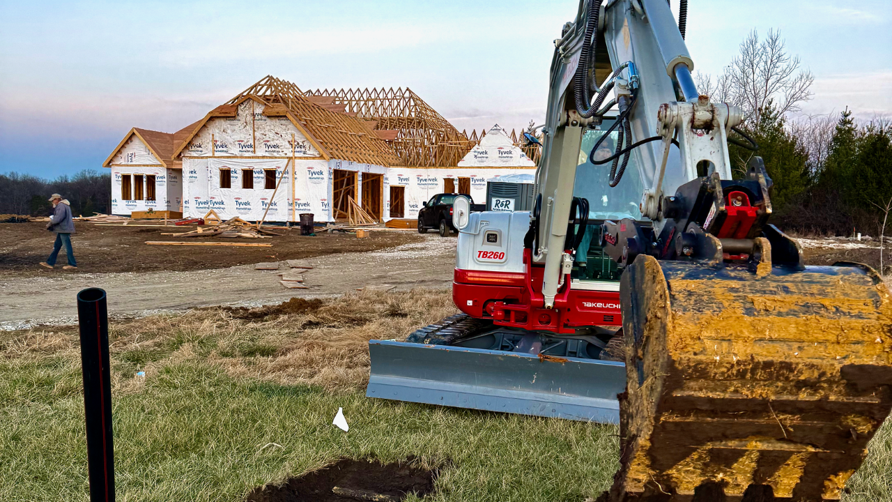 Construction site with a partially built house, a small excavator in the foreground, and a person walking nearby on a cloudy day.