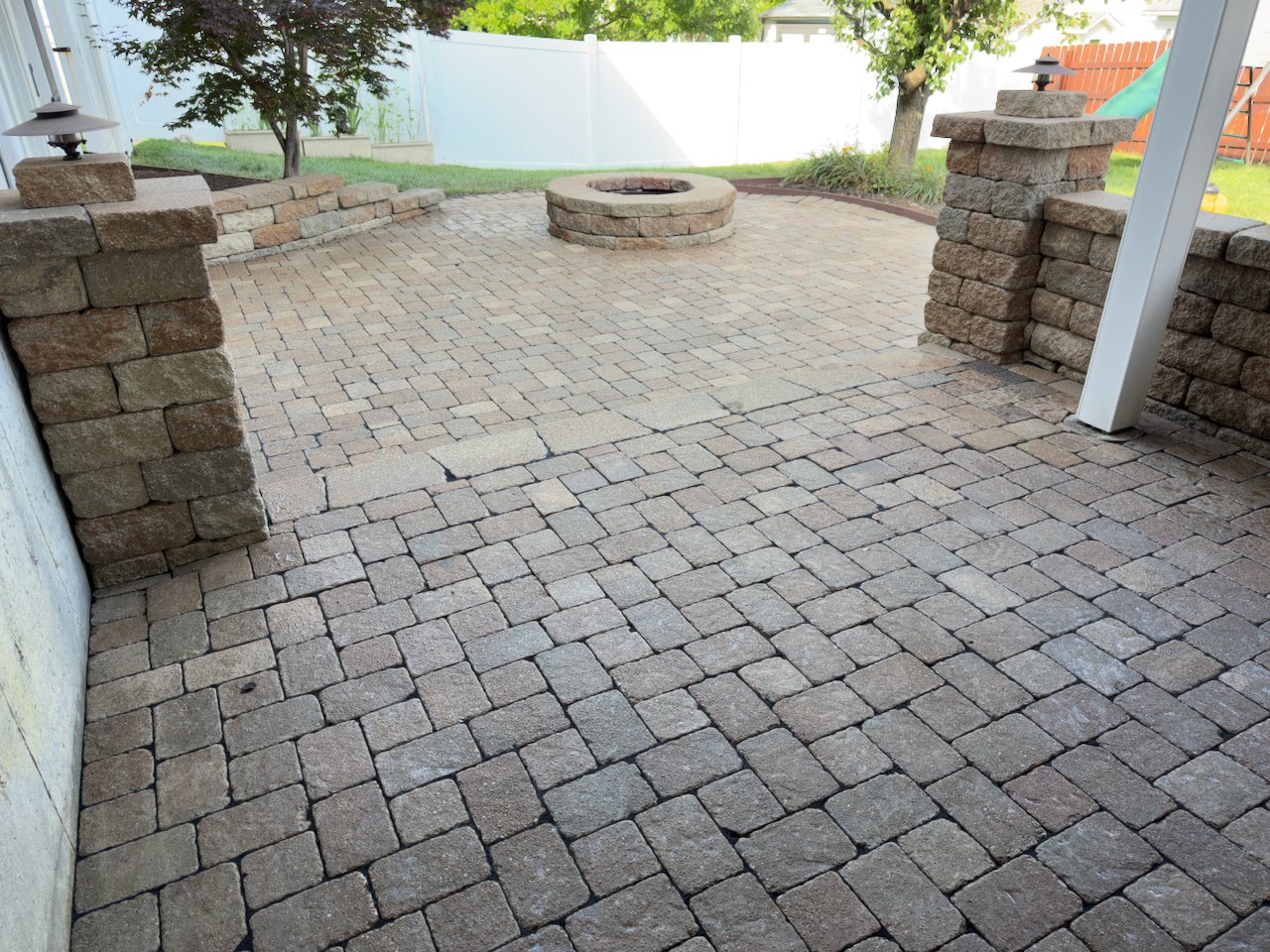 Backyard patio area with brick pavers, a fire pit, stone pillars with outdoor lights, and a green slide in the background.