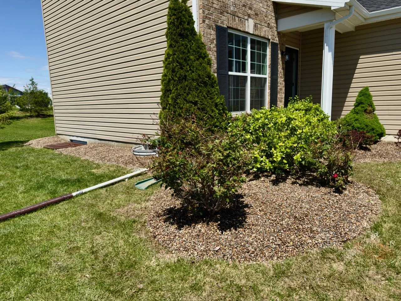 A suburban house with beige vinyl siding and a brick section around a window, surrounded by a landscaped yard with green grass, bushes, and small trees.