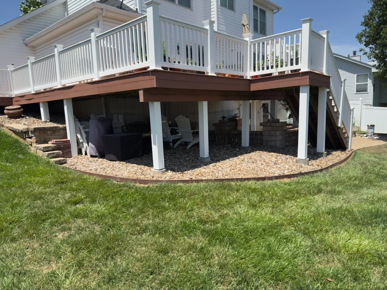 Backyard view of a raised deck with white railing, stairs, and covered patio area underneath with outdoor furniture and plants.