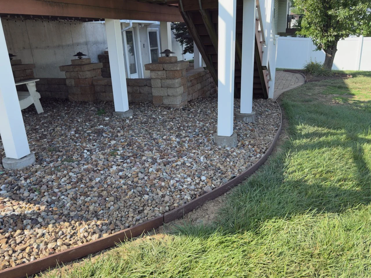 Backyard view showing a raised deck with stairs, supported by white posts on concrete blocks, over a bed of small multicolored rocks. There is grass and a curved border around the rocky area. A tree and a paved pathway are visible in the background.