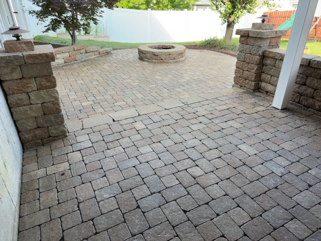 Backyard patio with brick pavers, brick and stone columns, a central fire pit, trees, and a white fence.