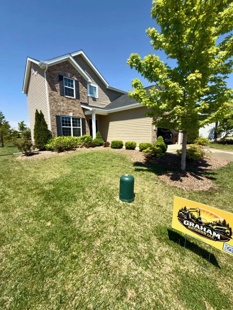 Front yard of a two-story house with beige siding and brick accents, a large green tree, well-kept bushes, and a lawn with a small sign that reads 'Graham Outdoor LLC' indicating landscaping or outdoor service.