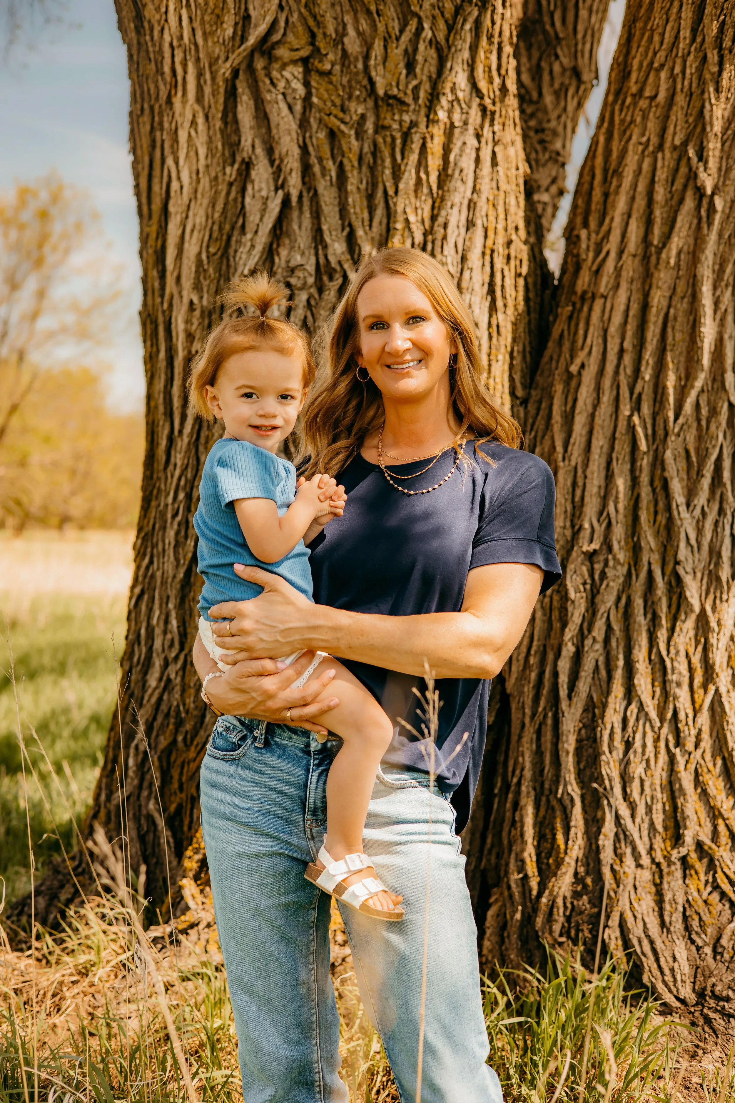 A woman with long hair wearing a dark blue shirt and light blue jeans holds a young girl with blonde hair tied up in a ponytail, wearing a blue shirt, in front of a large tree with textured bark in a grassy outdoor setting during daytime.