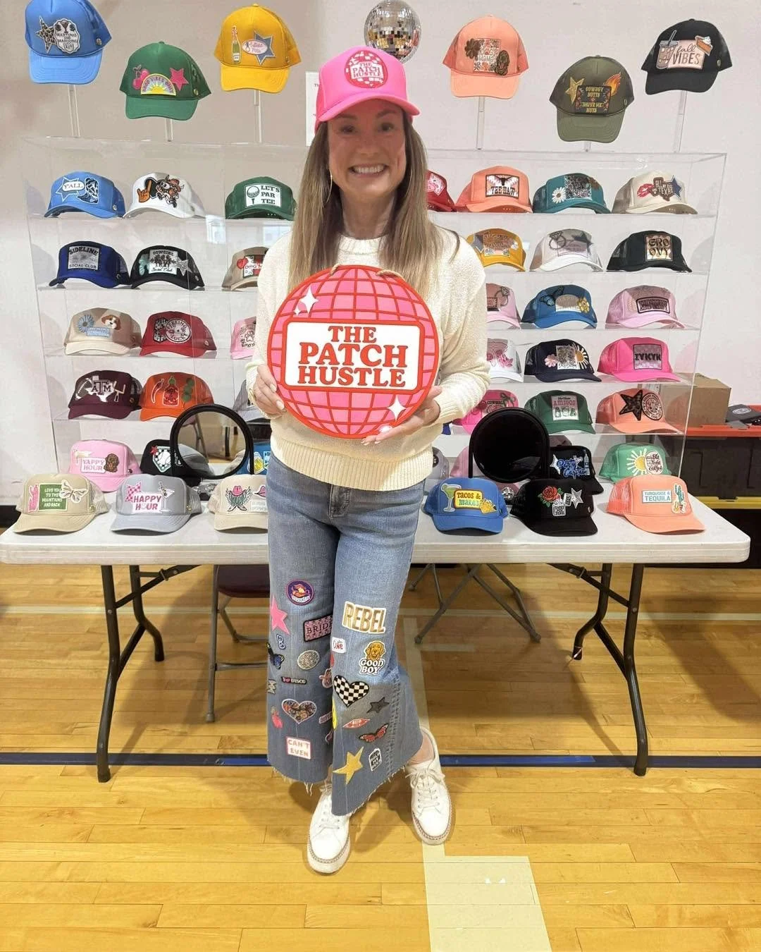 Woman in a sweater and jeans holding a pink round sign that says "The Patch Hustle," standing in front of a display of colorful hats with various patches on them.