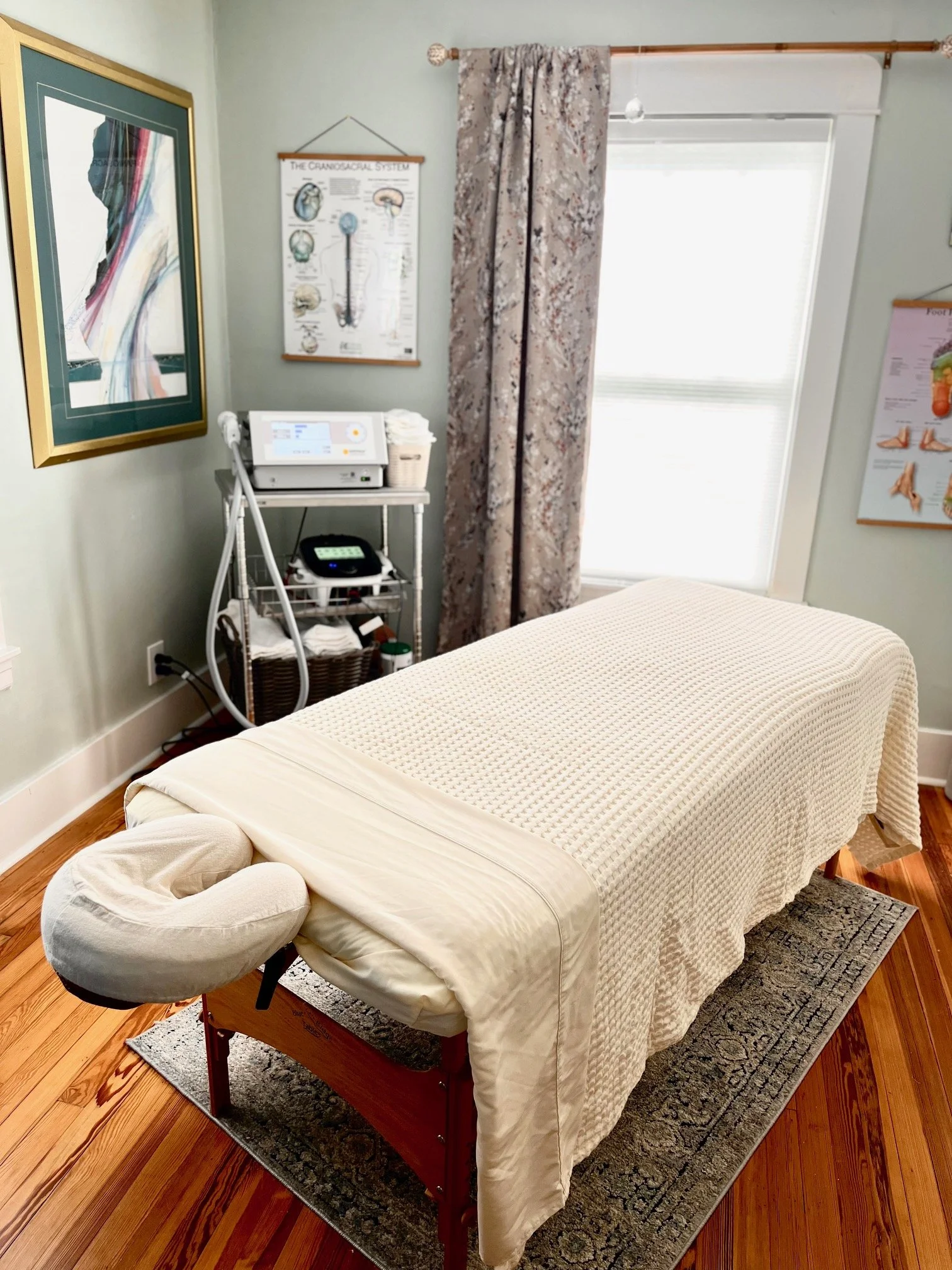 A massage table covered with a cream-colored quilt, with a cushioned face cradle at one end. The table is set on a dark rug on a wooden floor in a room with medical posters, curtains, and a window.