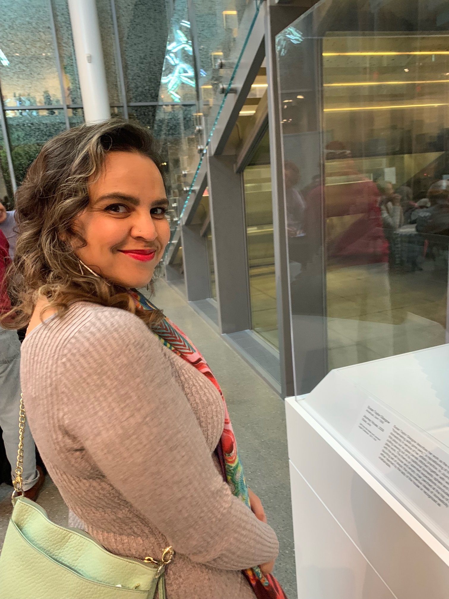 A woman with wavy hair and red lipstick standing at a display case in a modern indoor space, possibly a museum or gallery.