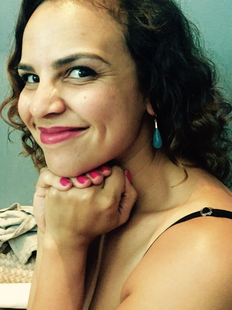 A woman with curly dark hair and pink lipstick smiles, resting her chin on her hand with nails painted pink, wearing a blue teardrop earring.