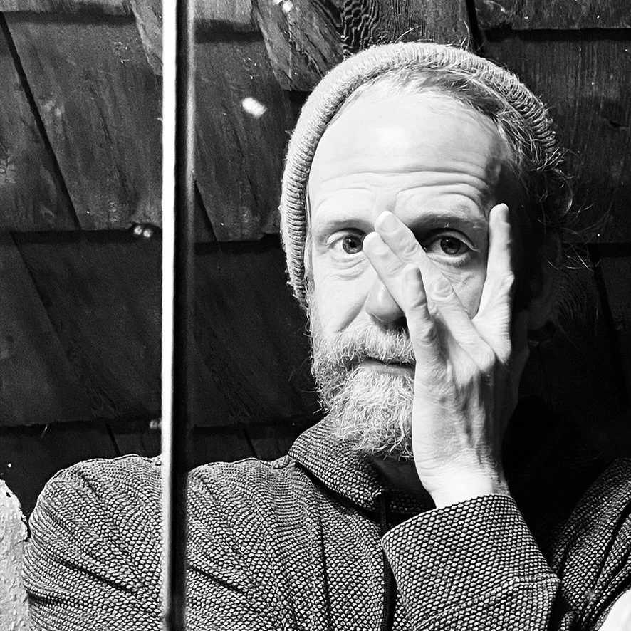 A black and white photo of a man with a beard and mustache, wearing a knit beanie and a textured jacket, holding his hand up over his eye making a gesture with two fingers, standing in front of a wooden wall.
