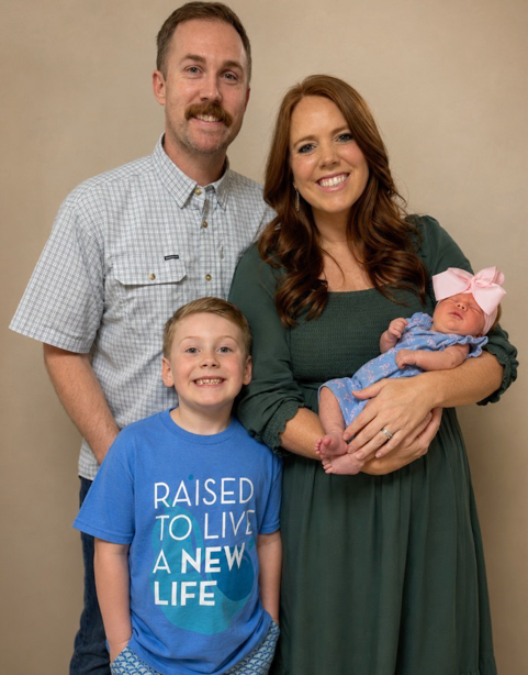 A family of four, including a woman holding a newborn with a pink bow, a smiling young boy wearing a blue T-shirt with the text "Raised to Live a New Life," a man, and the baby, posing against a beige background.