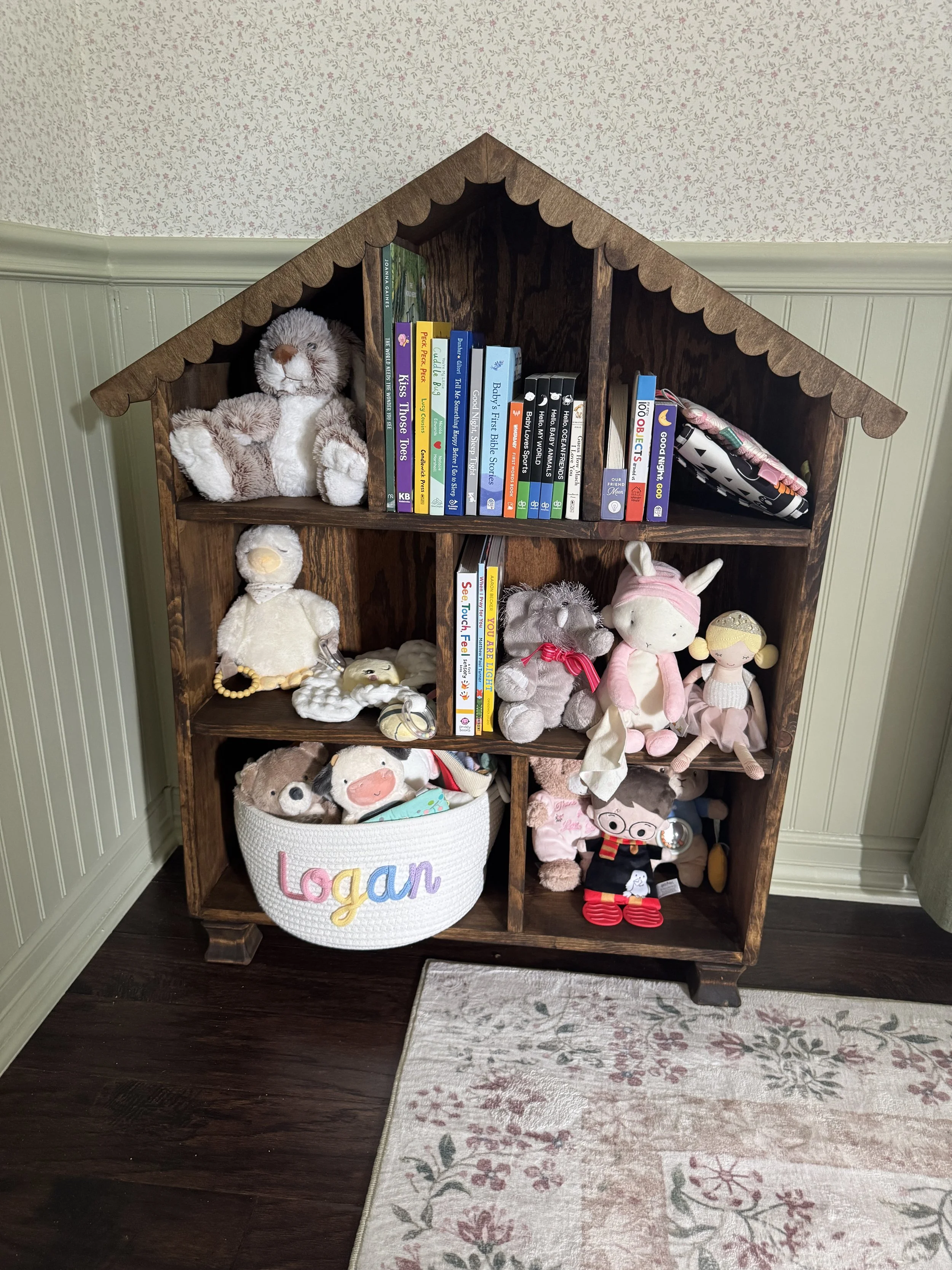 A wooden bookshelf with a scalloped roof filled with children's books and stuffed animals, including a plush lion and a pink stuffed bunny, beside a basket with the name "Logan" embroidered on it, set on a dark hardwood floor and a floral area rug.