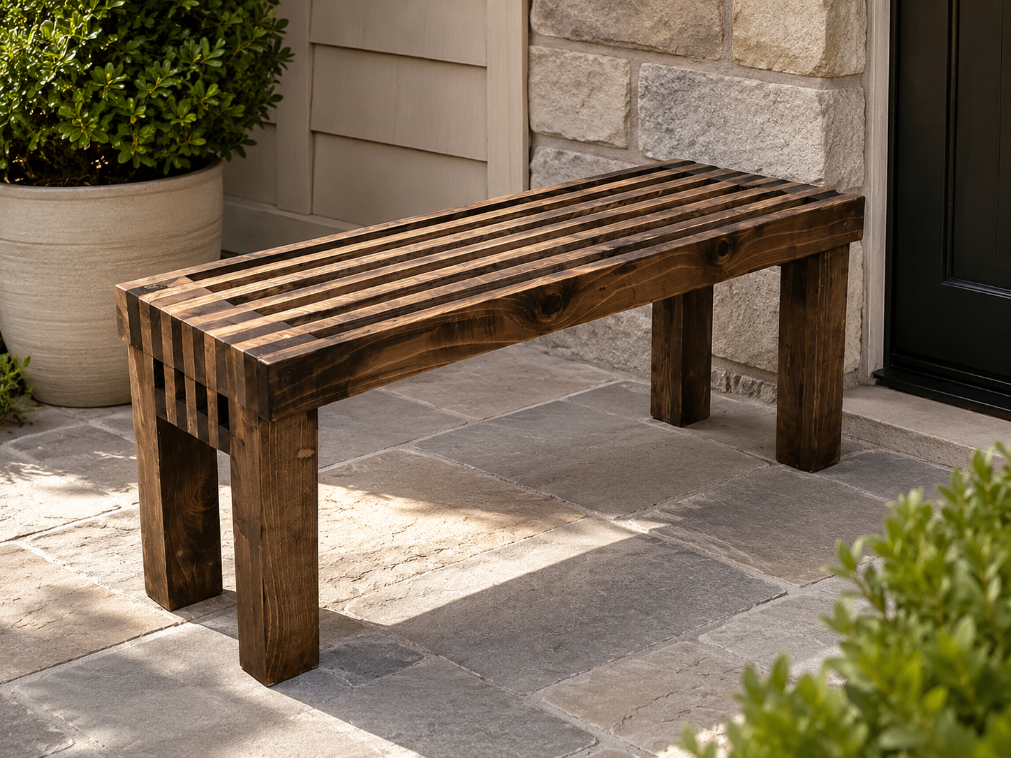 A wooden slatted outdoor bench on a stone patio next to a potted shrub and a house wall.