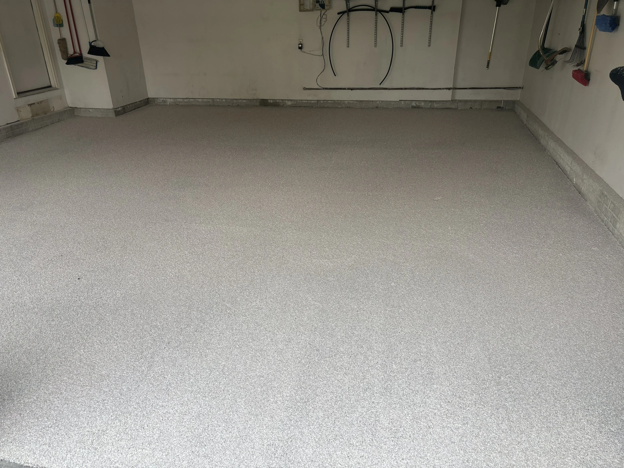 Unfinished garage with new epoxy coated floor, garden tools hanging on the wall, and a broom and dustpan leaning against the wall.