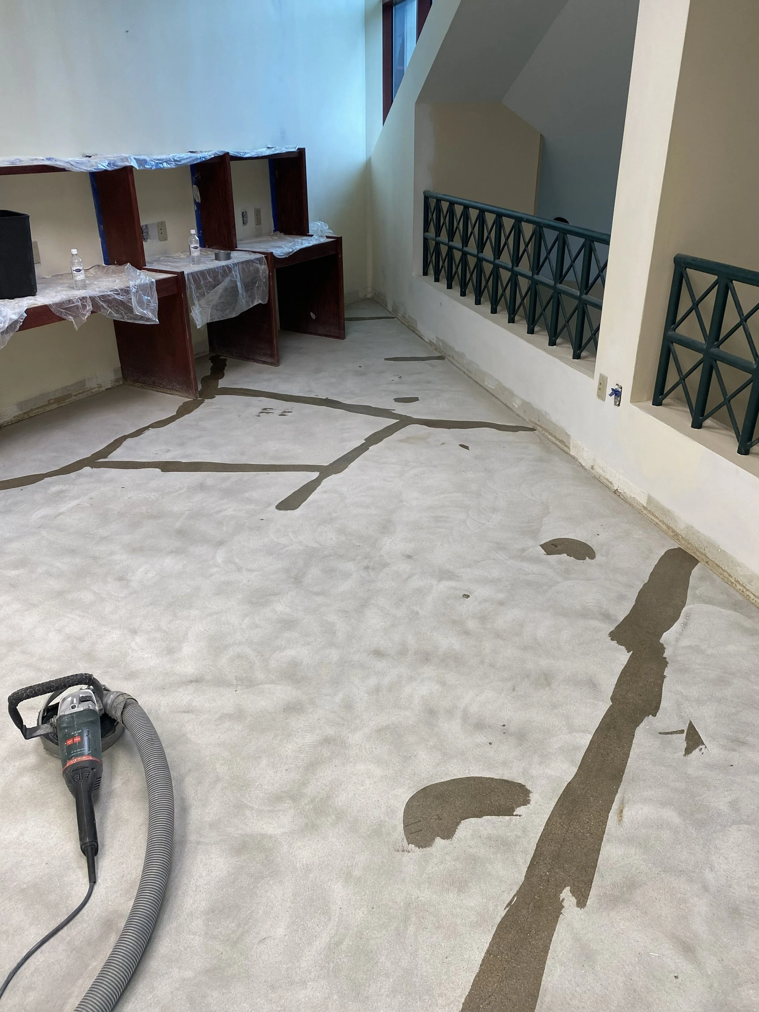Floor with a cracks filled with a dark gray sealant or epoxy, a portable vacuum cleaner or shop vac on the floor, part of a staircase with a green railing, and over a row of empty wooden desks covered with plastic sheeting, with some bottles of water and electrical outlets on the wall.