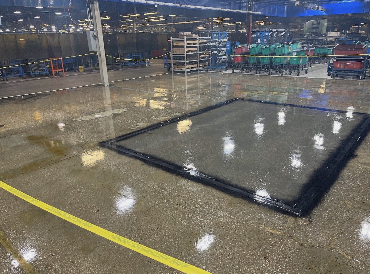 Empty retail or warehouse floor with a large square hatch marked with a black border, surrounded by wet epoxy and a yellow outline. Overhead lights reflect on the wet surface, with shelves and carts visible in the background.