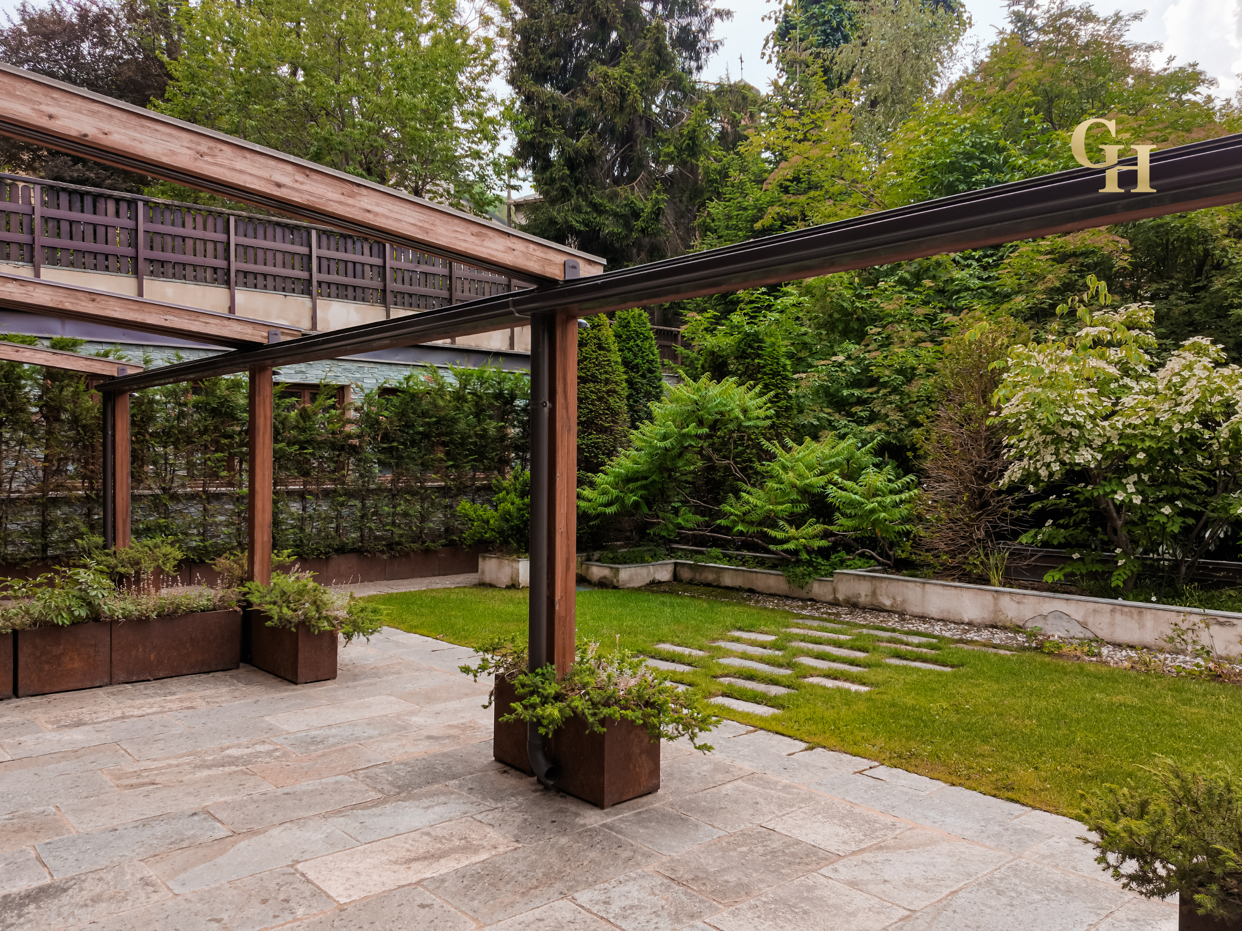 A backyard garden with a patio, potted plants, a grassy lawn, trees, and a wooden fence with a trellis.
