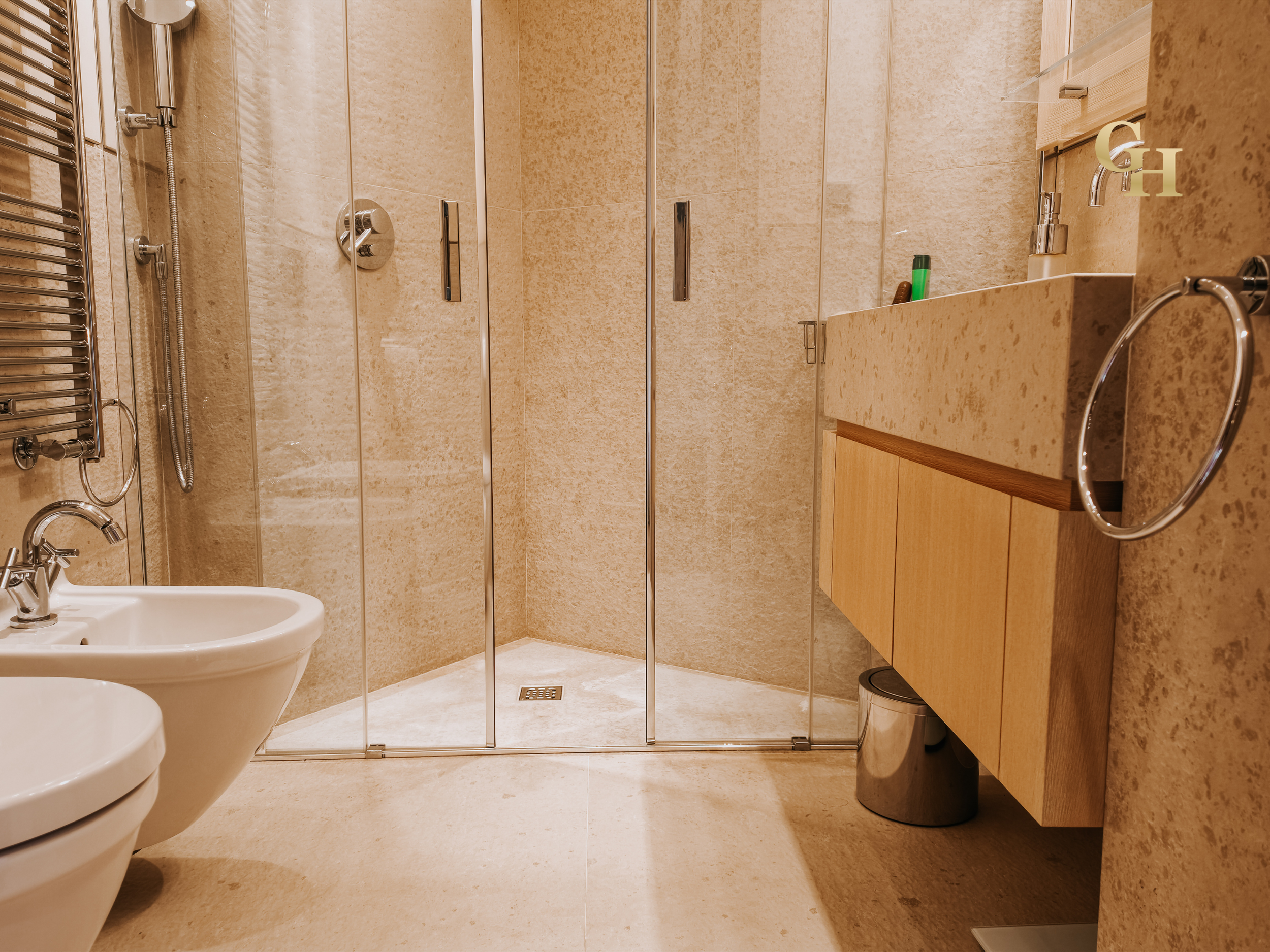 A beige-tiled bathroom with a glass shower enclosure, a bidet, a wooden vanity with a sink, a mirror, a towel ring, a small waste bin, and a small shelf above the sink.
