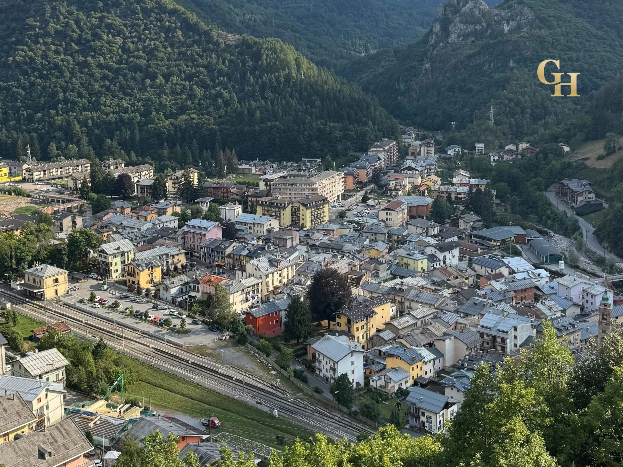 Aerial view of a small town nestled in lush green mountains with residential buildings, streets, and railway tracks.