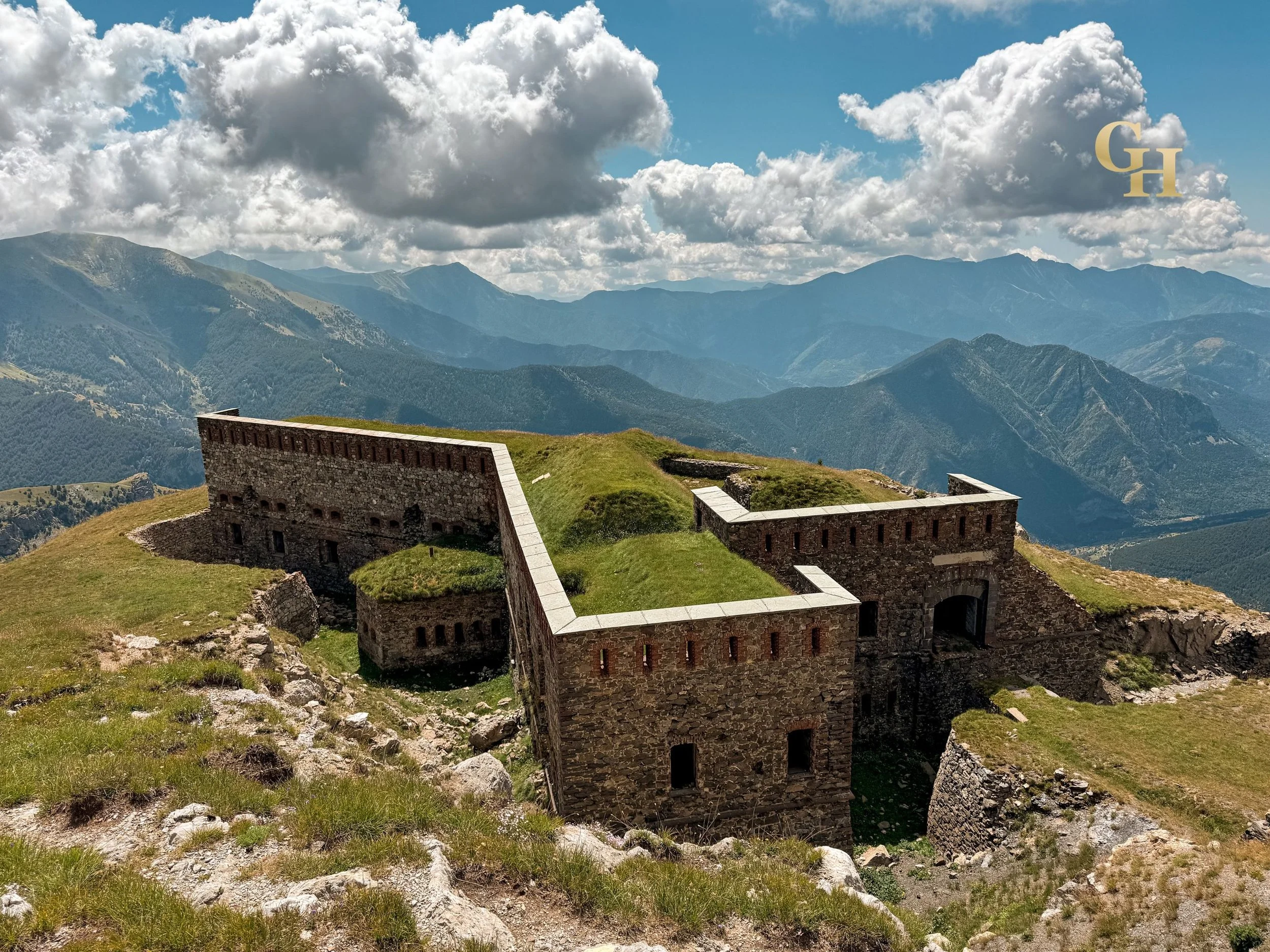 Ancient stone fortress on a grassy hill with mountains in the background under a partly cloudy sky.