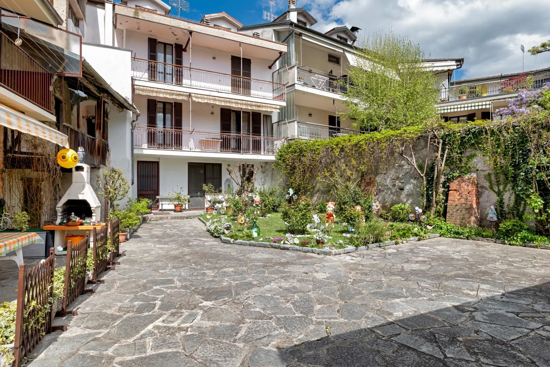 A residential courtyard with a stone-paved ground, small garden with plants and decorative flowers, surrounded by multi-story apartment buildings with balconies, some with awnings and plants. There's a chimney and outdoor furniture visible, and the s