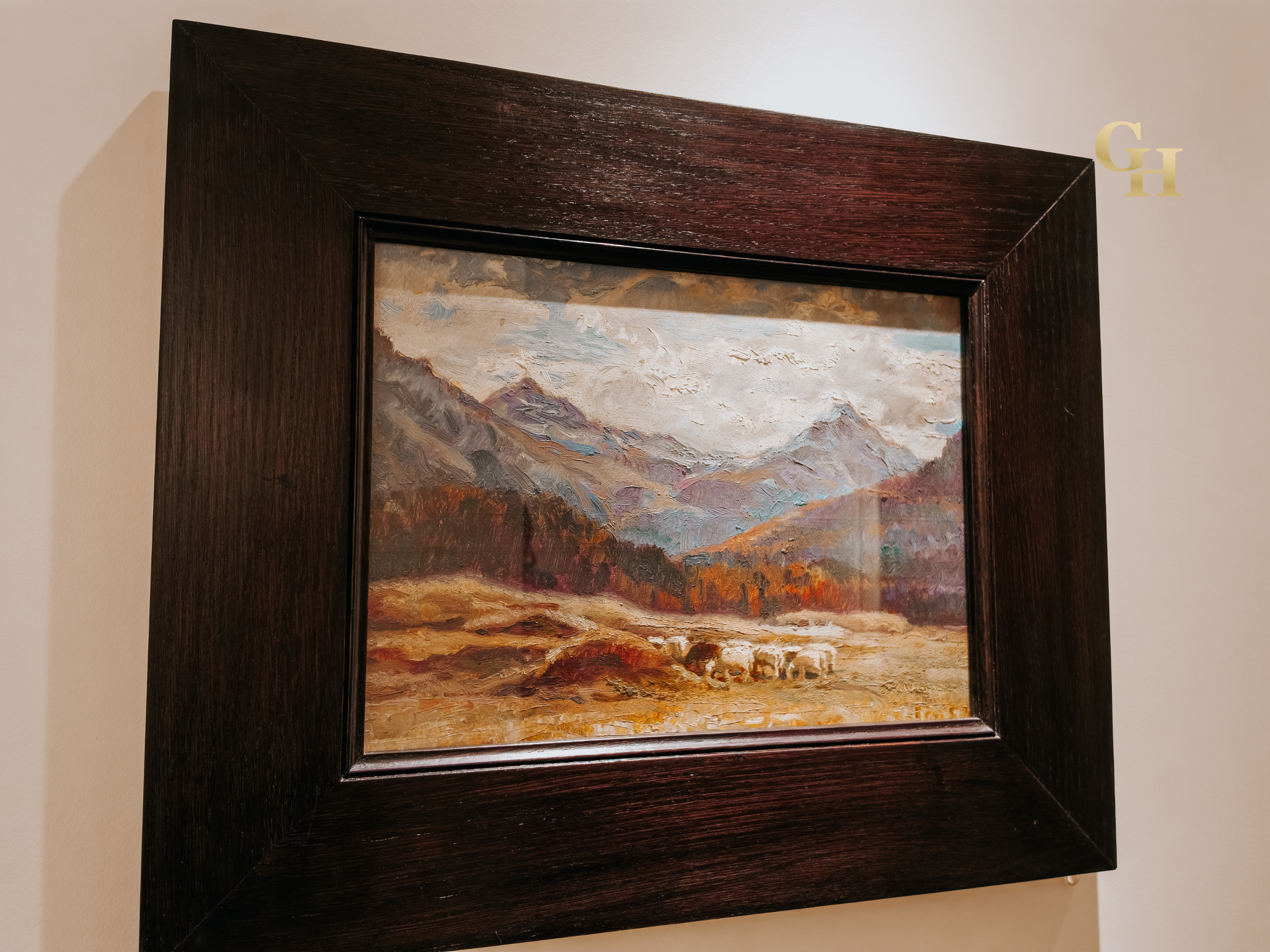 Framed landscape painting with mountains, sky, and trees, mounted on a beige wall.