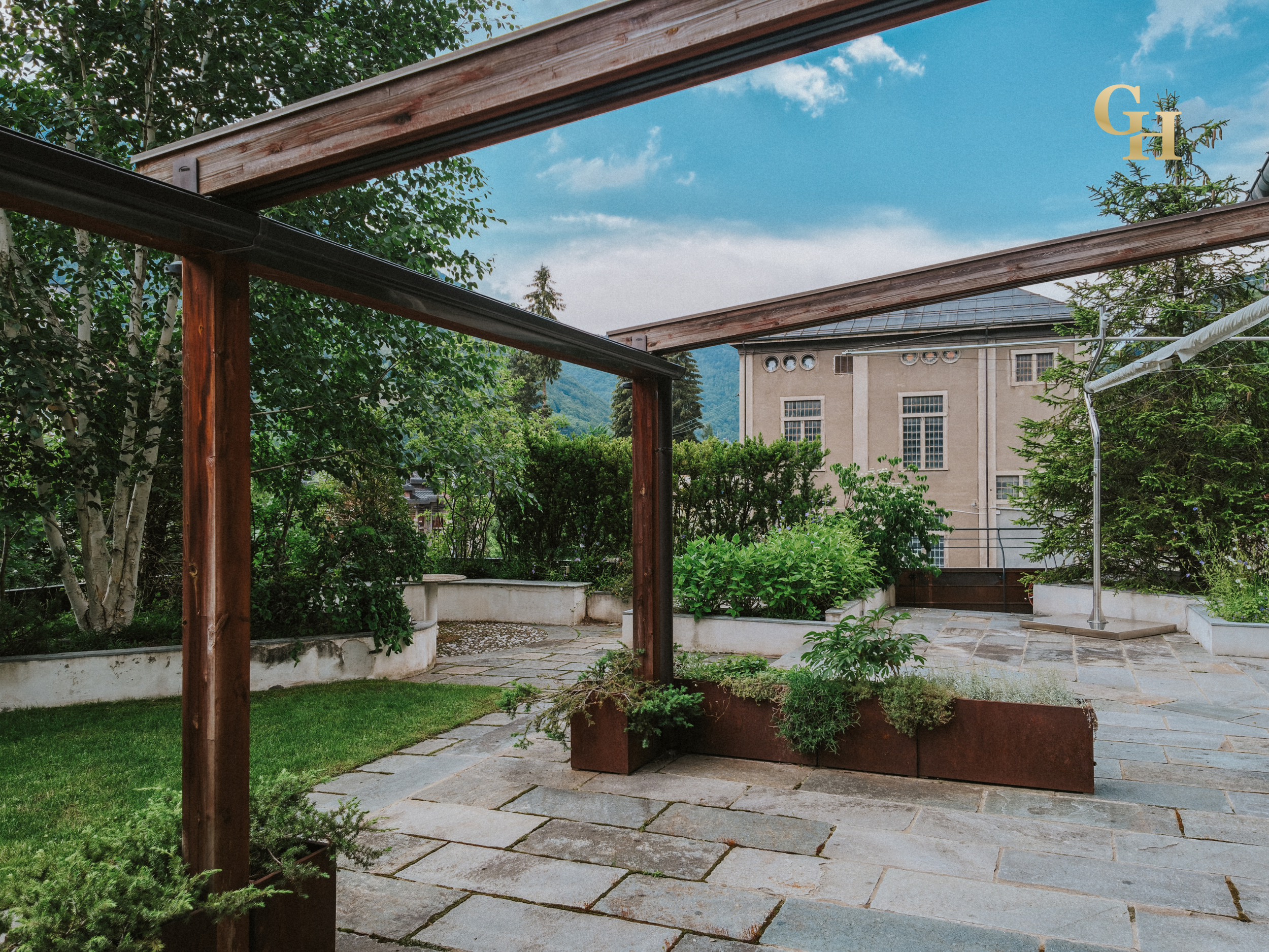Backyard patio with stone pavers, green grass, trees, and a building with multiple windows in the background. There is a wooden and metal structure extending over part of the patio.