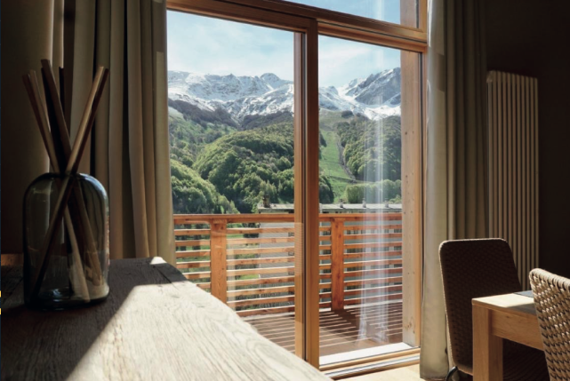 Room with sliding glass door leading to a balcony overlooking mountains with snow and green trees.