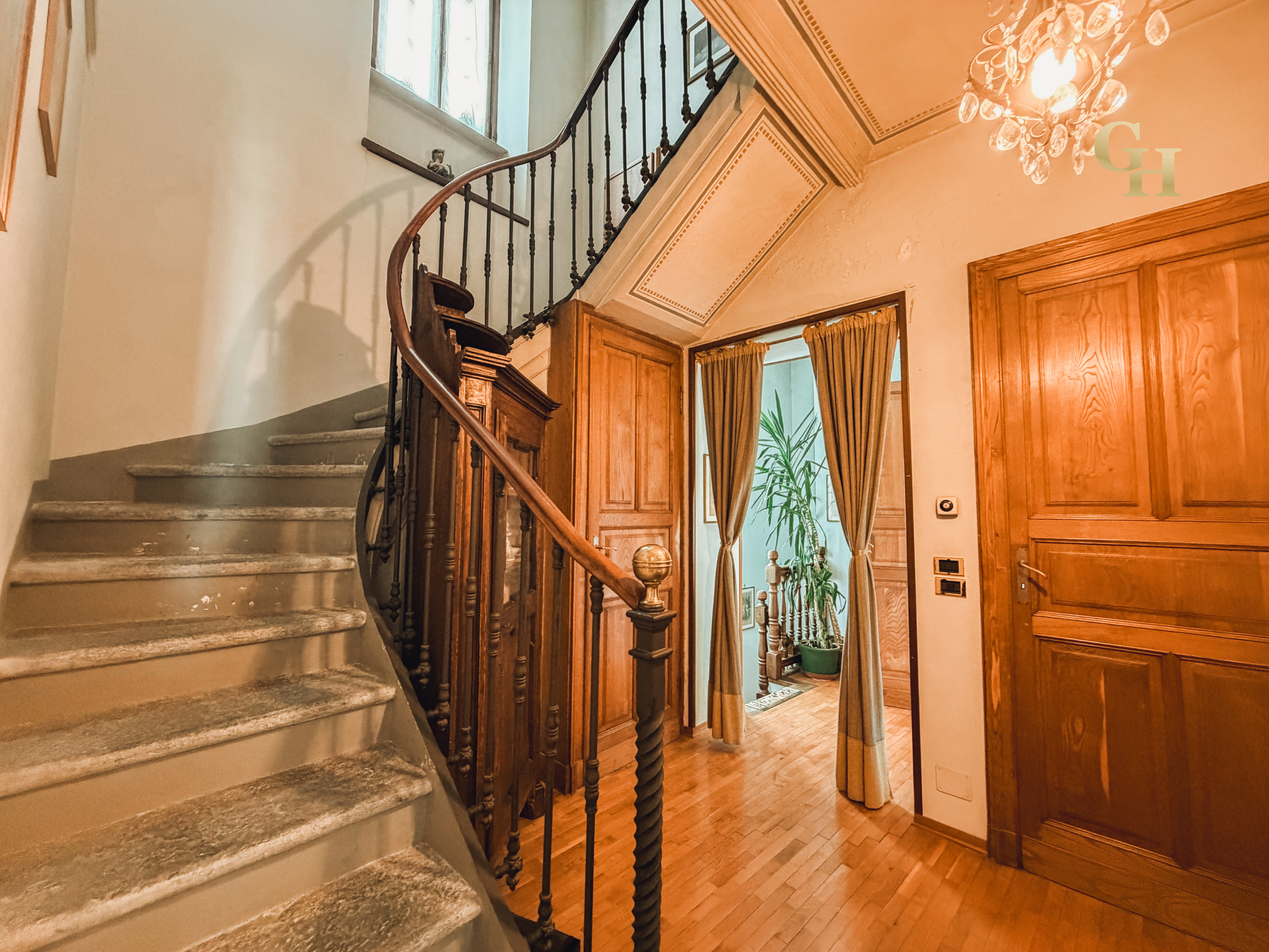 Interior view of a home featuring a staircase with worn stone steps, a black metal and wooden handrail, and a wooden doorway with curtains. There is a window at the top of the staircase with a small statue on the sill, and a chandelier hanging from t