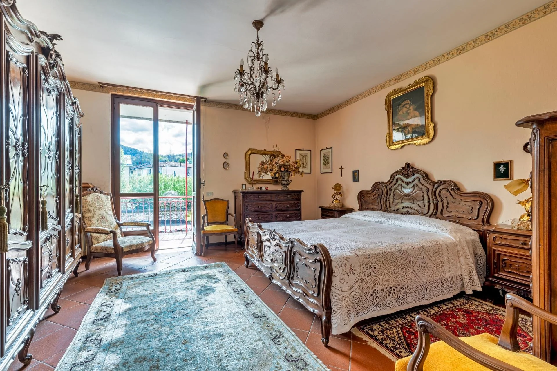 A vintage bedroom with a wooden bed, matching furniture, and ornate decorations. There is a large window with a view of mountains and greenery outside, a chandelier hanging from the ceiling, and various framed pictures on the walls.