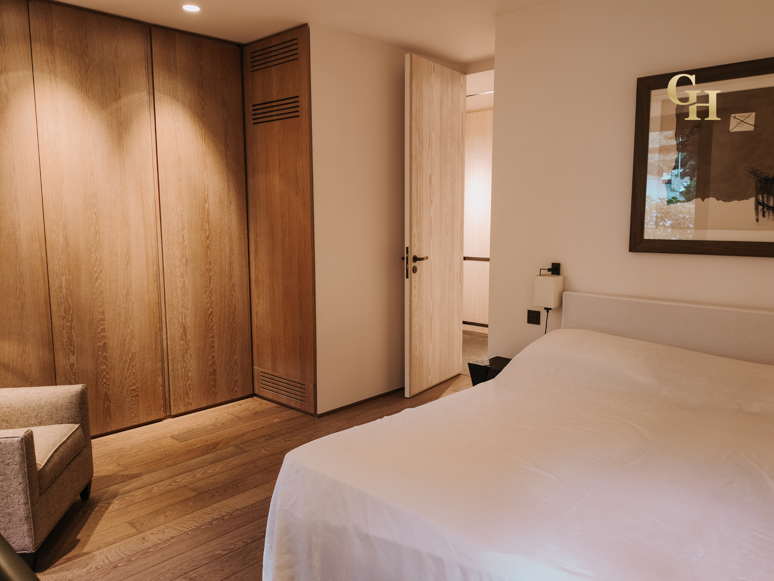 Interior of a modern bedroom with wooden flooring and a large bed with white bedding. There is a wooden wardrobe on the left side, a partially open door leading to another room, and a framed abstract art piece hanging on the wall. The room has minima