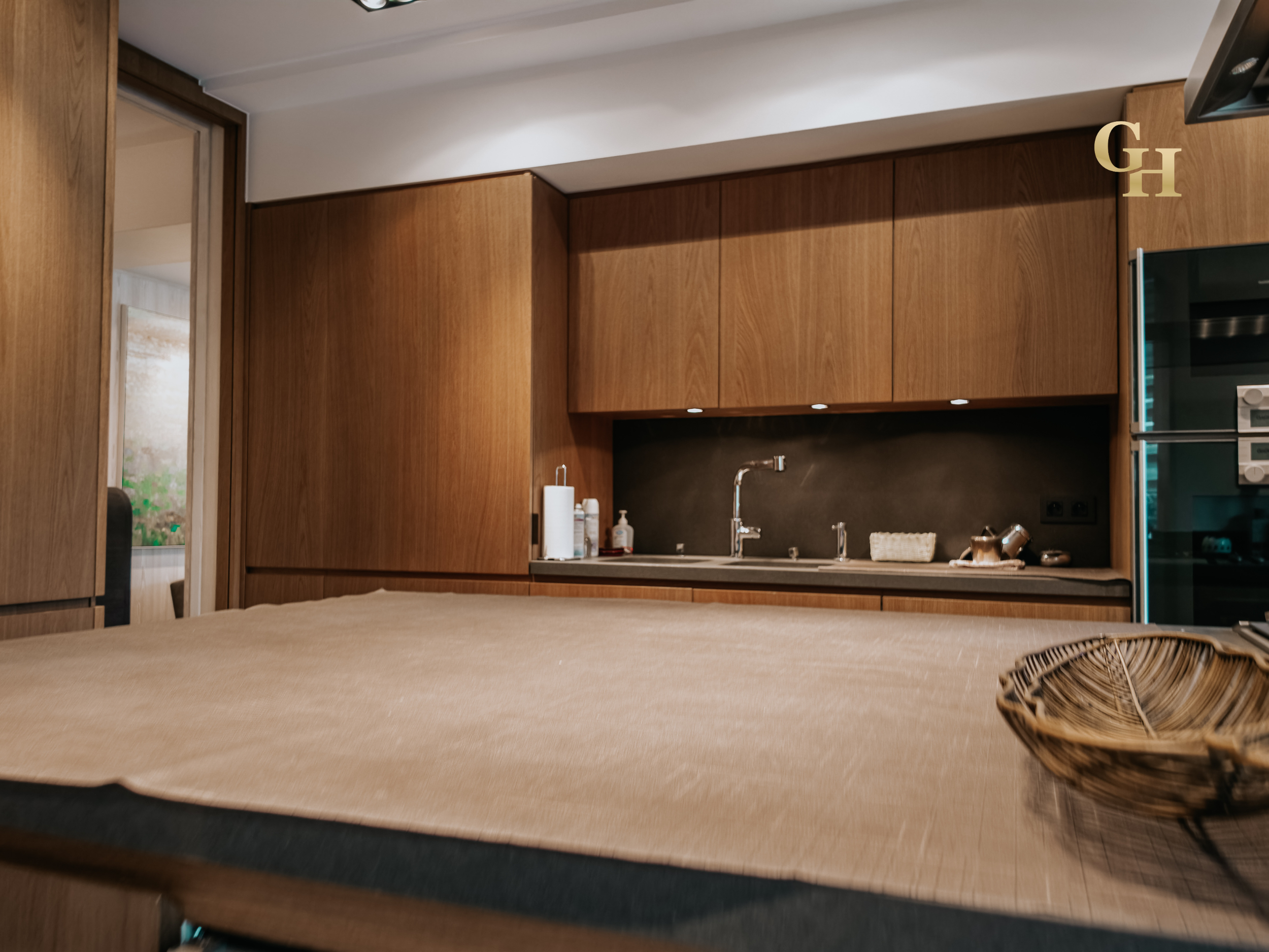 A kitchen with wooden cabinets, a sink, and countertop, seen through a mirror with a reflection of a window. A wicker basket is faded in the foreground.