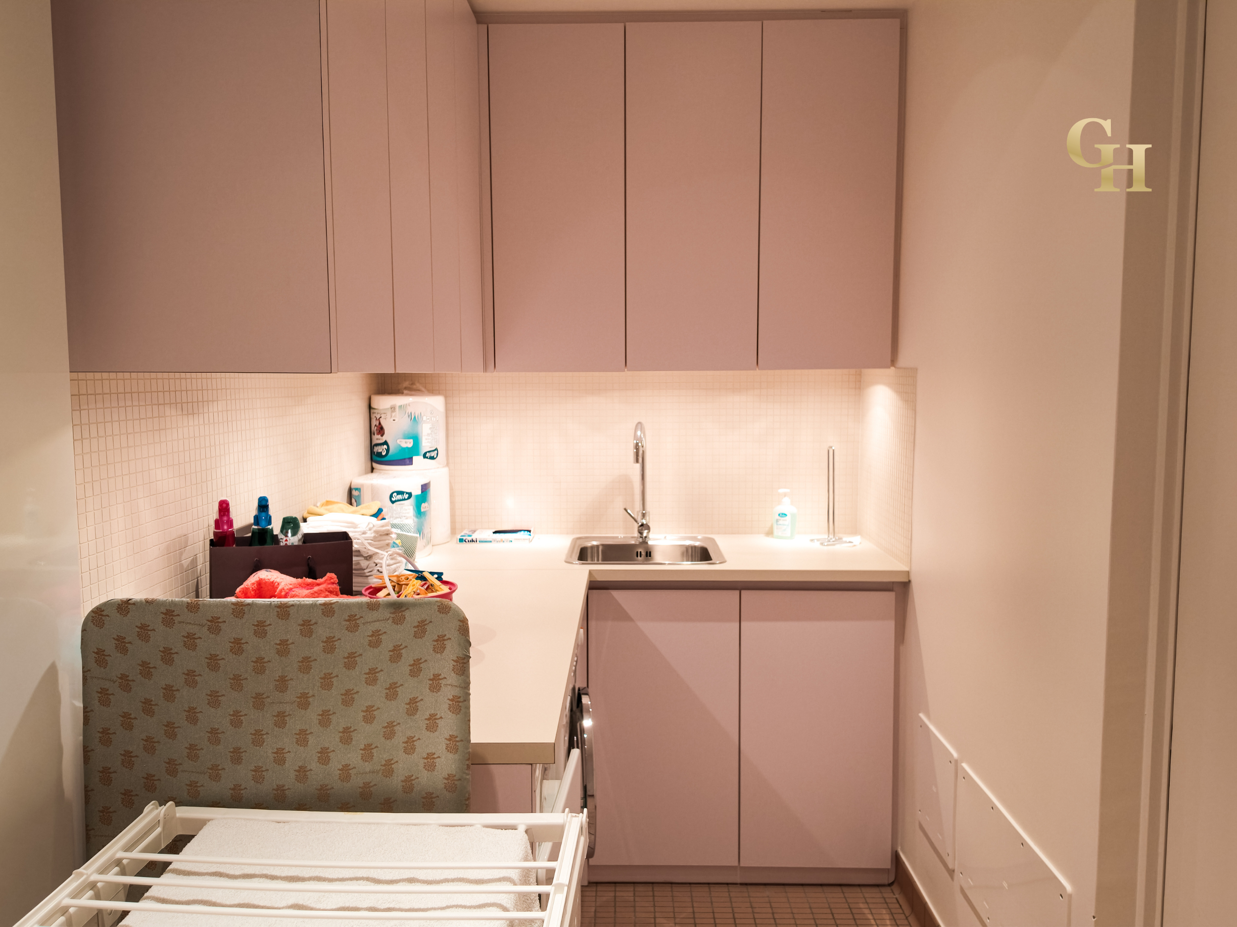 A small kitchen with pink cabinets, a white countertop, and a stainless steel sink. Items on the counter include a tissue box, baby wipes, and cleaning supplies. There is a patterned chair with a towel draped over it in the foreground. The wall behin