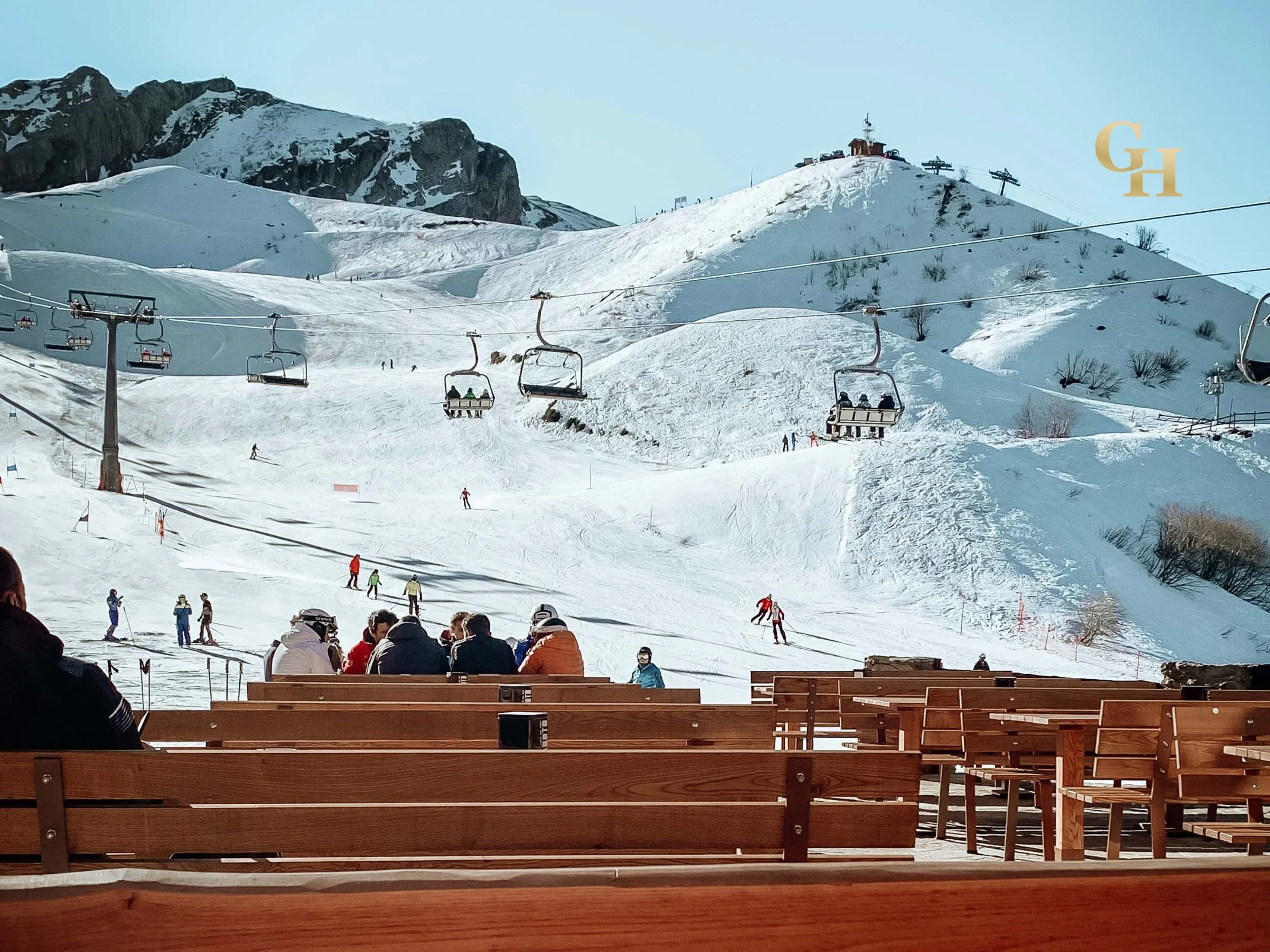 People sitting on wooden benches at a ski resort, overlooking snowy slopes with skiers and snowboarders, ski lifts, and mountain terrain in the background. Riserva Bianca Slopes, Maritime Alps, Piedmont, Italy.