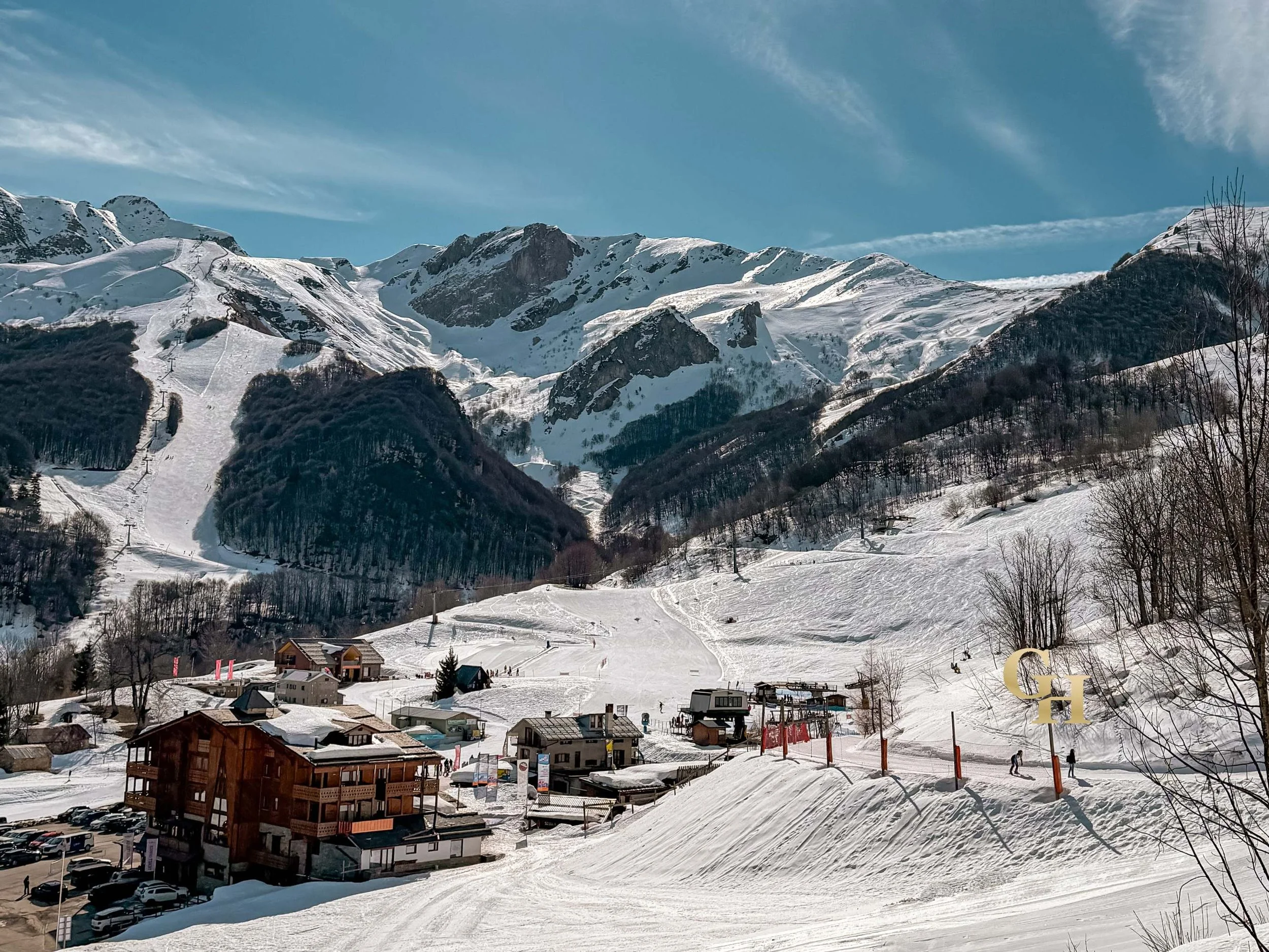 Snow-covered mountains with ski slopes and ski lifts, a small village at the base with buildings, trees, and parked cars, under a partly cloudy blue sky.