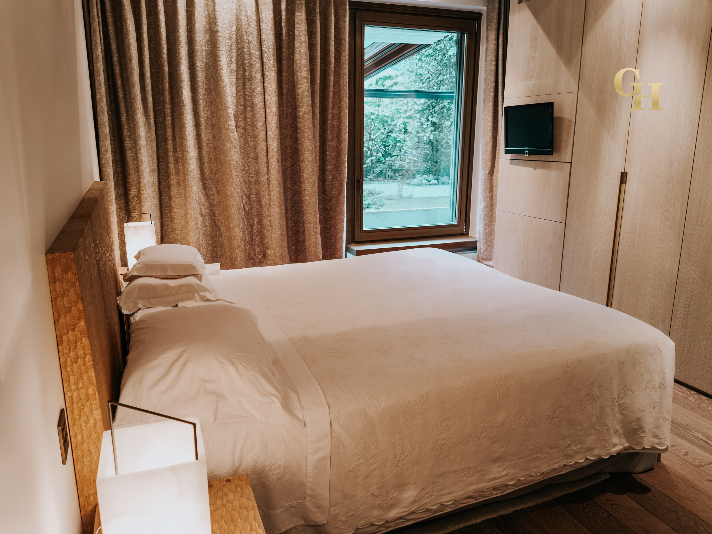 A cozy hotel room with a bed, a nightstand with a lamp, beige curtains, a window looking out to greenery, a wall-mounted TV, and a wooden wardrobe.
