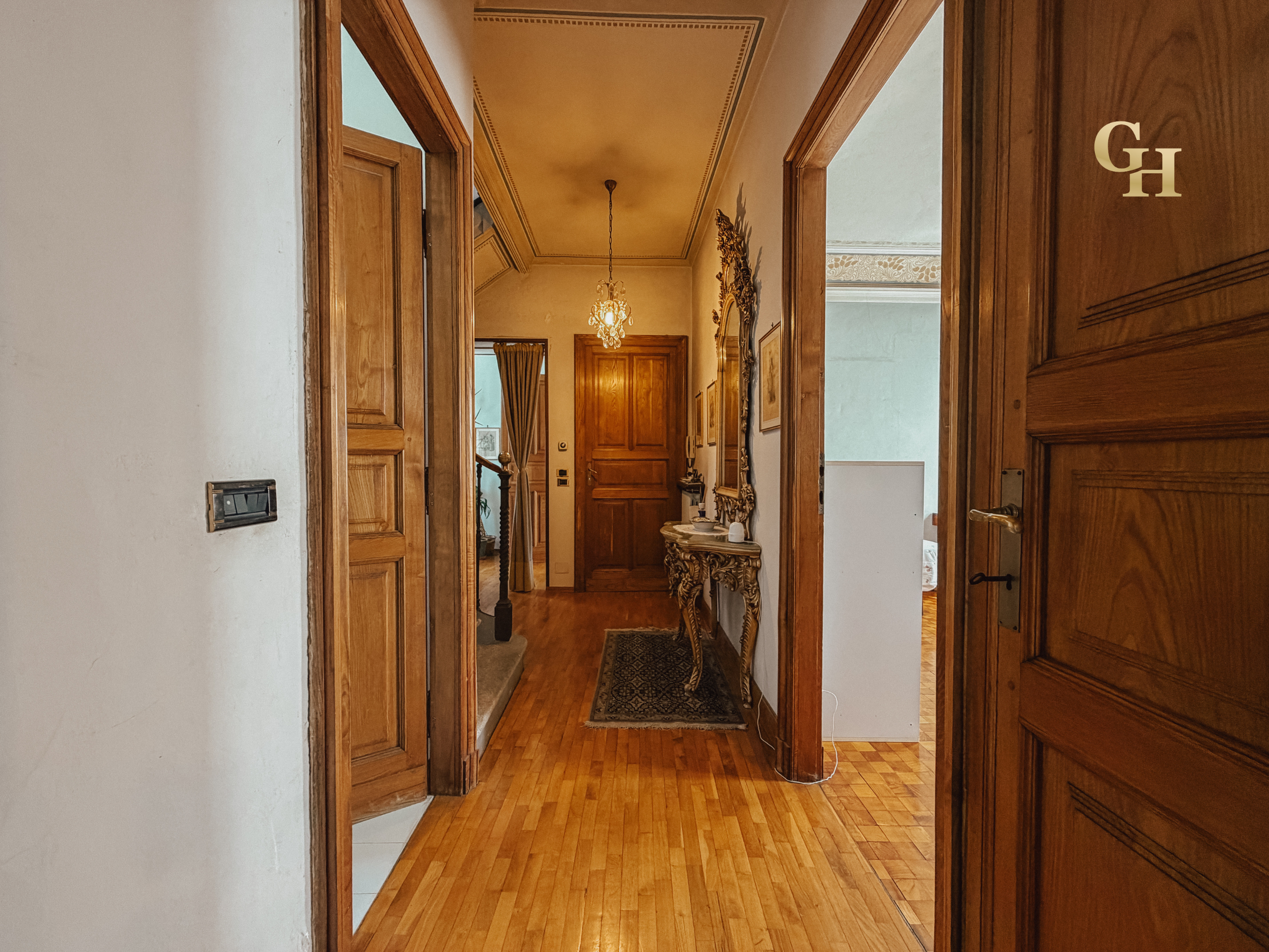 View of a hallway with light wood floors and doors, a chandelier hanging from the ceiling, and a mirror with ornate gold frame on the wall.