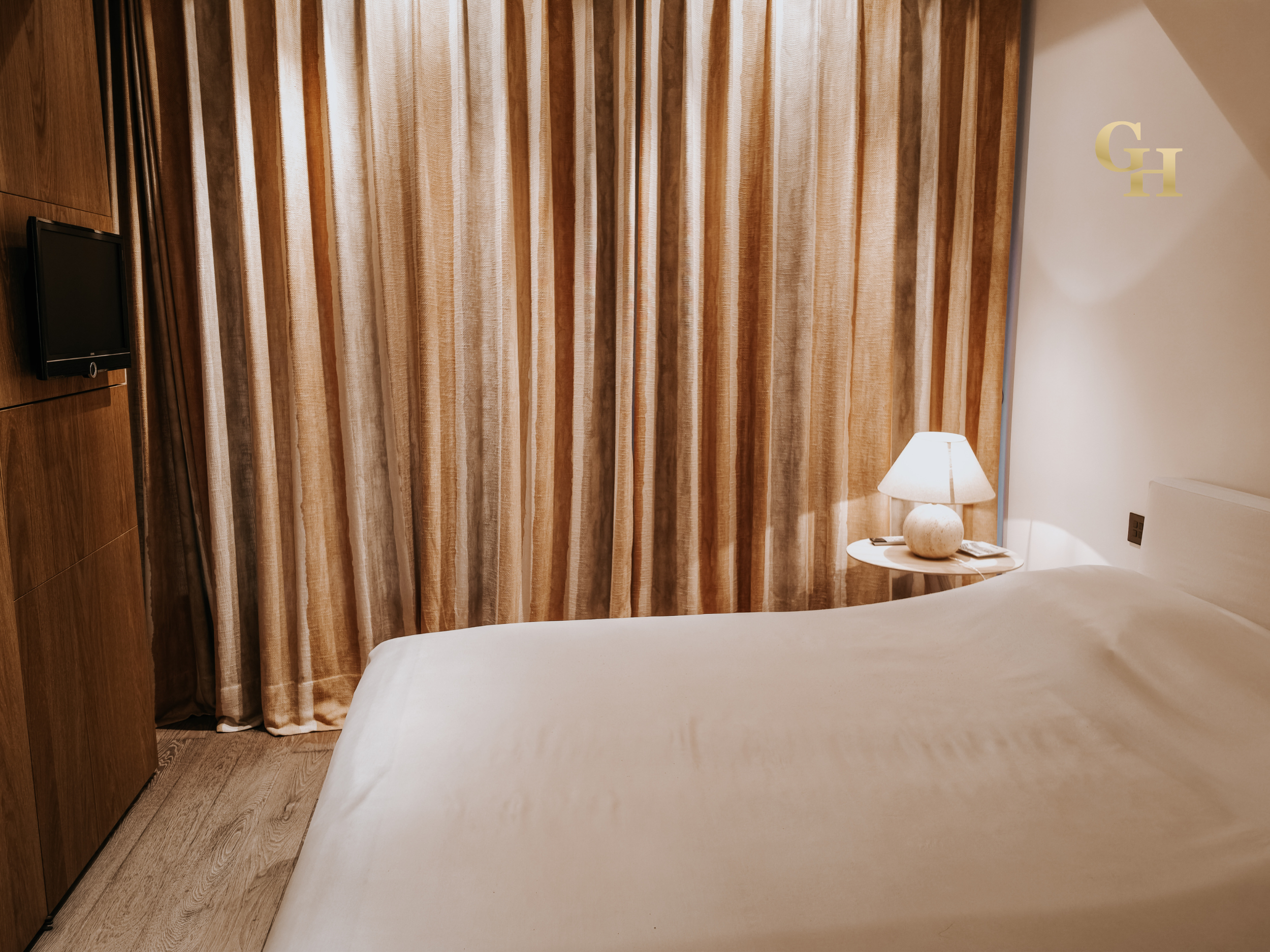 A corner of a hotel room with a bed, a nightstand with a lamp, curtain, small television, and wooden wall paneling.