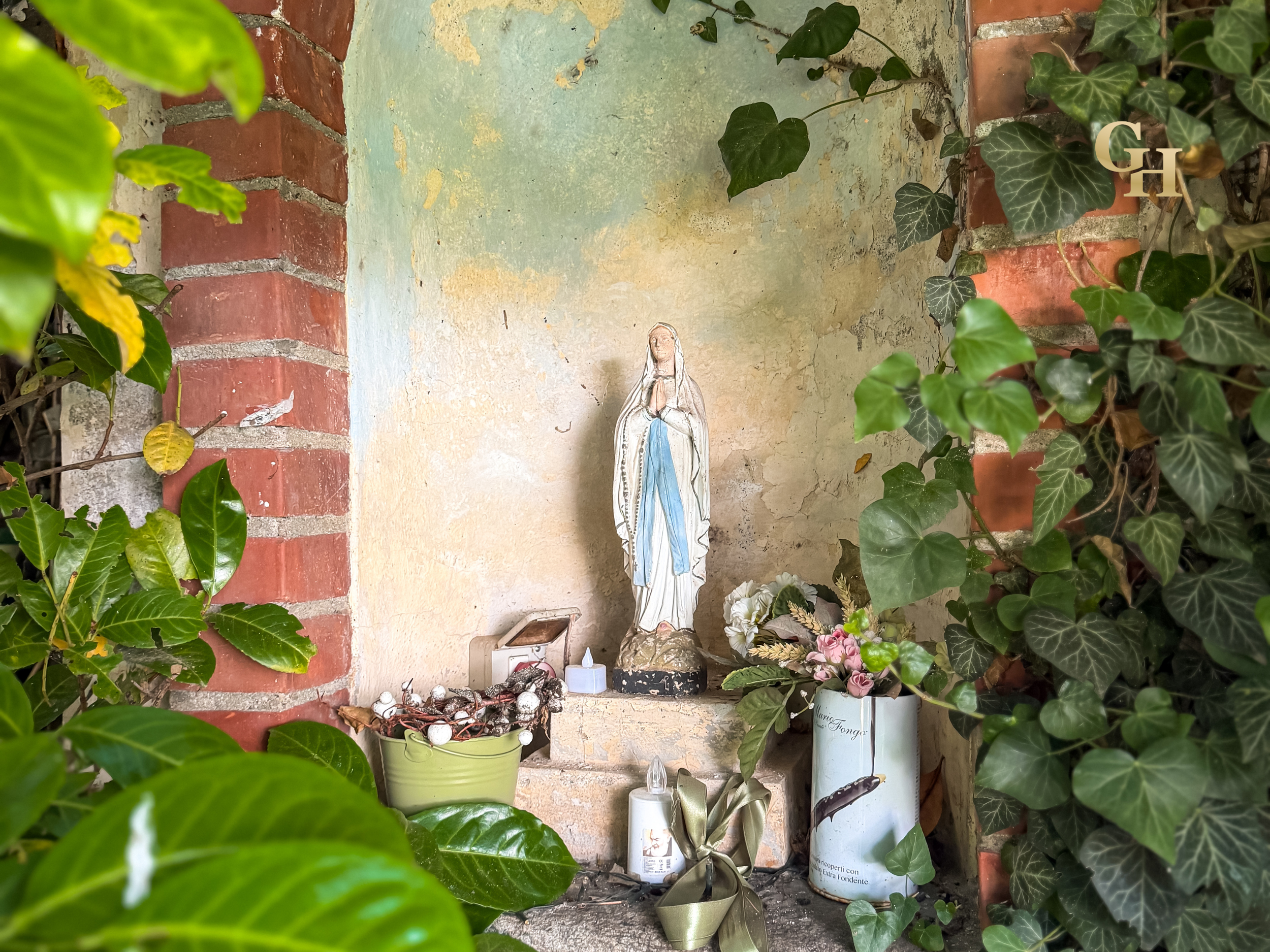 An outdoor altar with religious statues and plants, including ivy and other greenery, against a brick and painted wall background.
