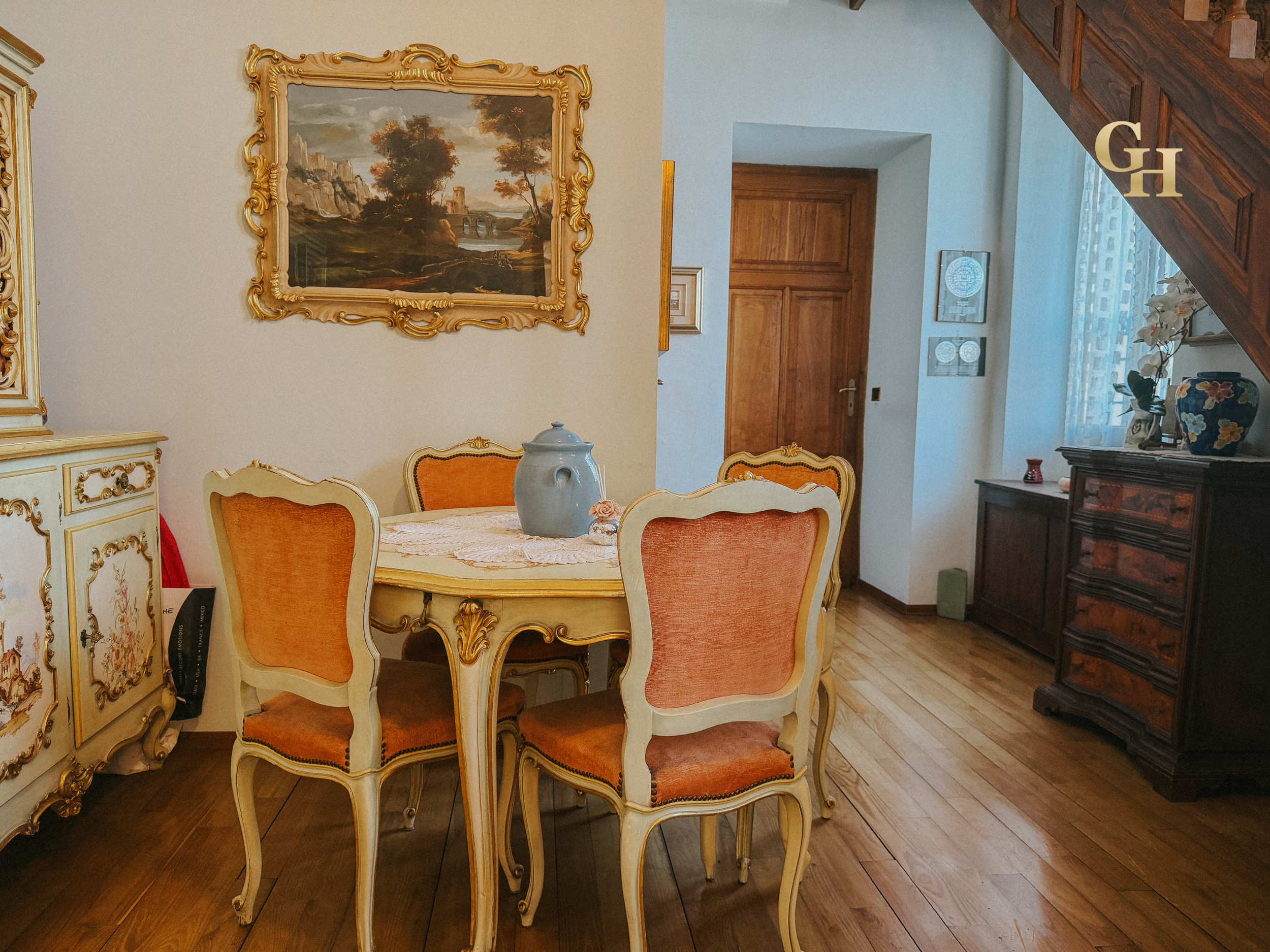 Interior of a dining room with a round table, upholstered chairs, a large landscape painting with a gold ornate frame on the wall, and a wooden staircase.