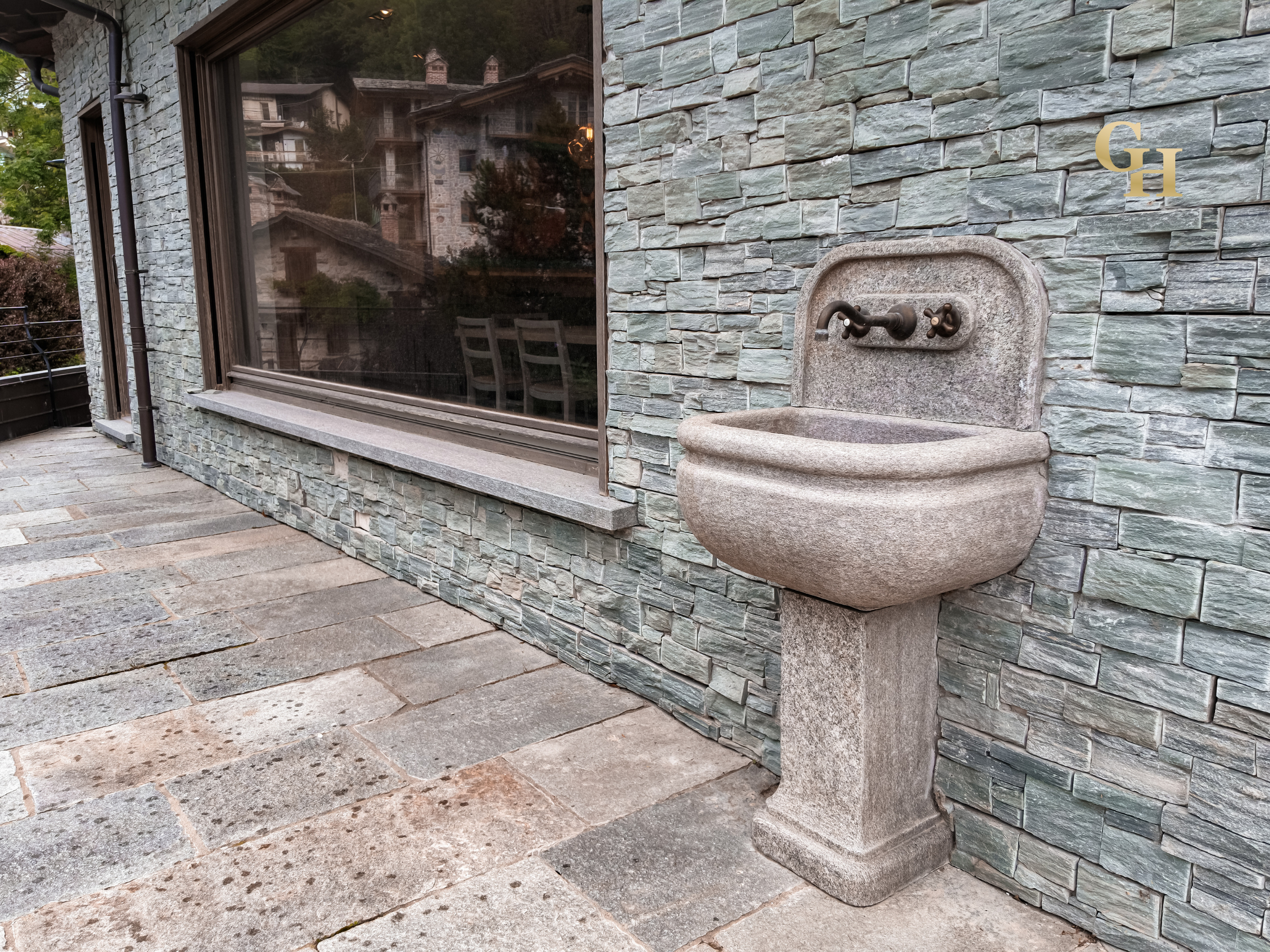 A stone wall exterior of a house with a large window and an outdoor stone sink attached to the wall.