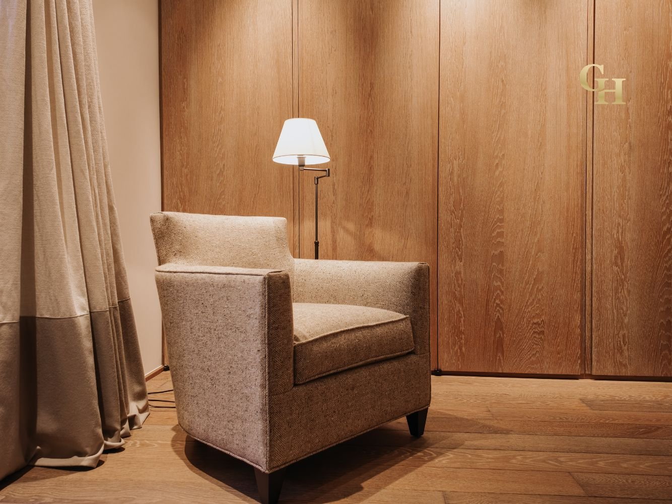 A beige upholstered armchair next to a floor lamp with a white shade on a wooden floor and wooden partition.