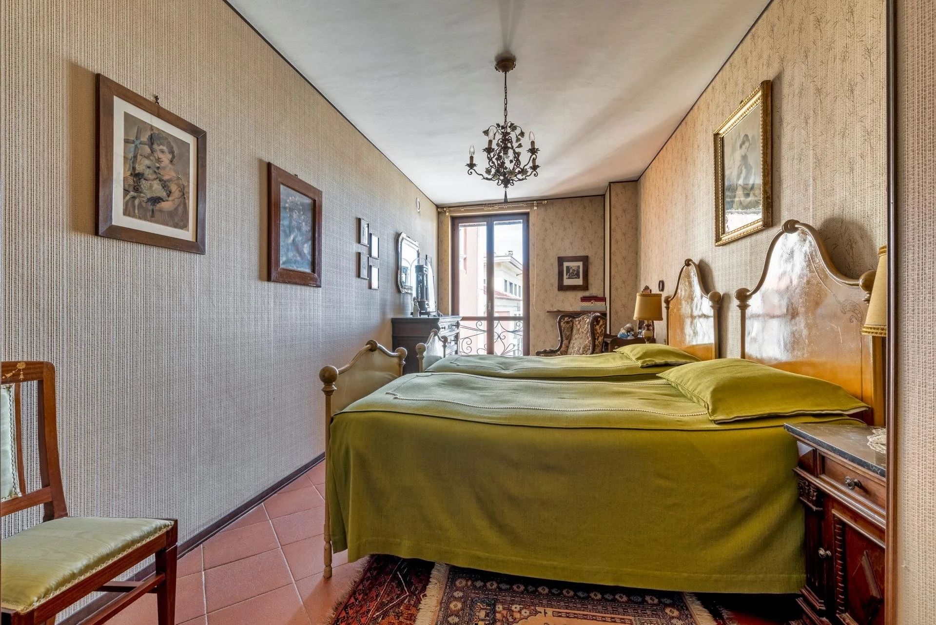 A bedroom with two twin beds with yellow-green bedding, wooden headboards, and matching nightstands. There are several framed pictures on the beige textured walls, a small table with a lampshade, a vintage chair near a window, and a chandelier hangin